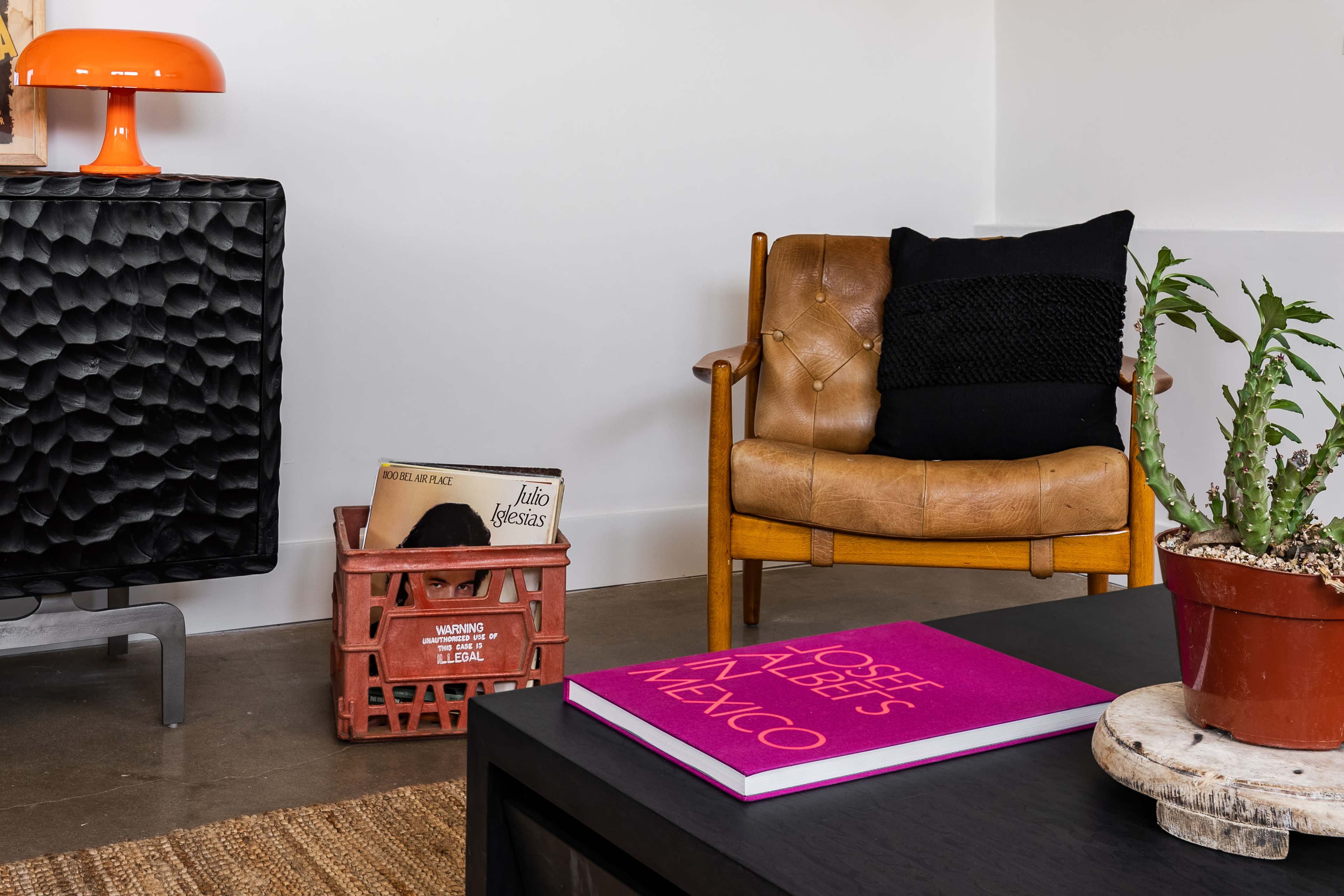 A stylish living room featuring a leather armchair, a patterned rug, a coffee table with a bright pink book, a basket of vinyl records, and a potted cactus.