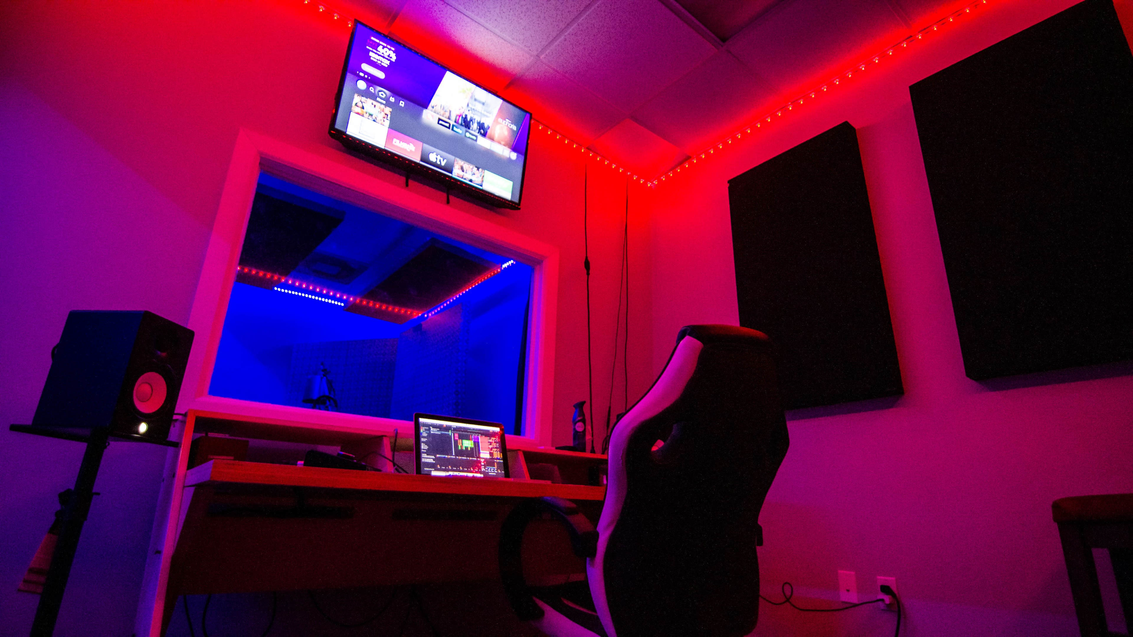 A recording studio features a desk with a computer and an ergonomic chair, illuminated by red and blue LED lighting, with soundproof panels on the walls and a TV mounted above.