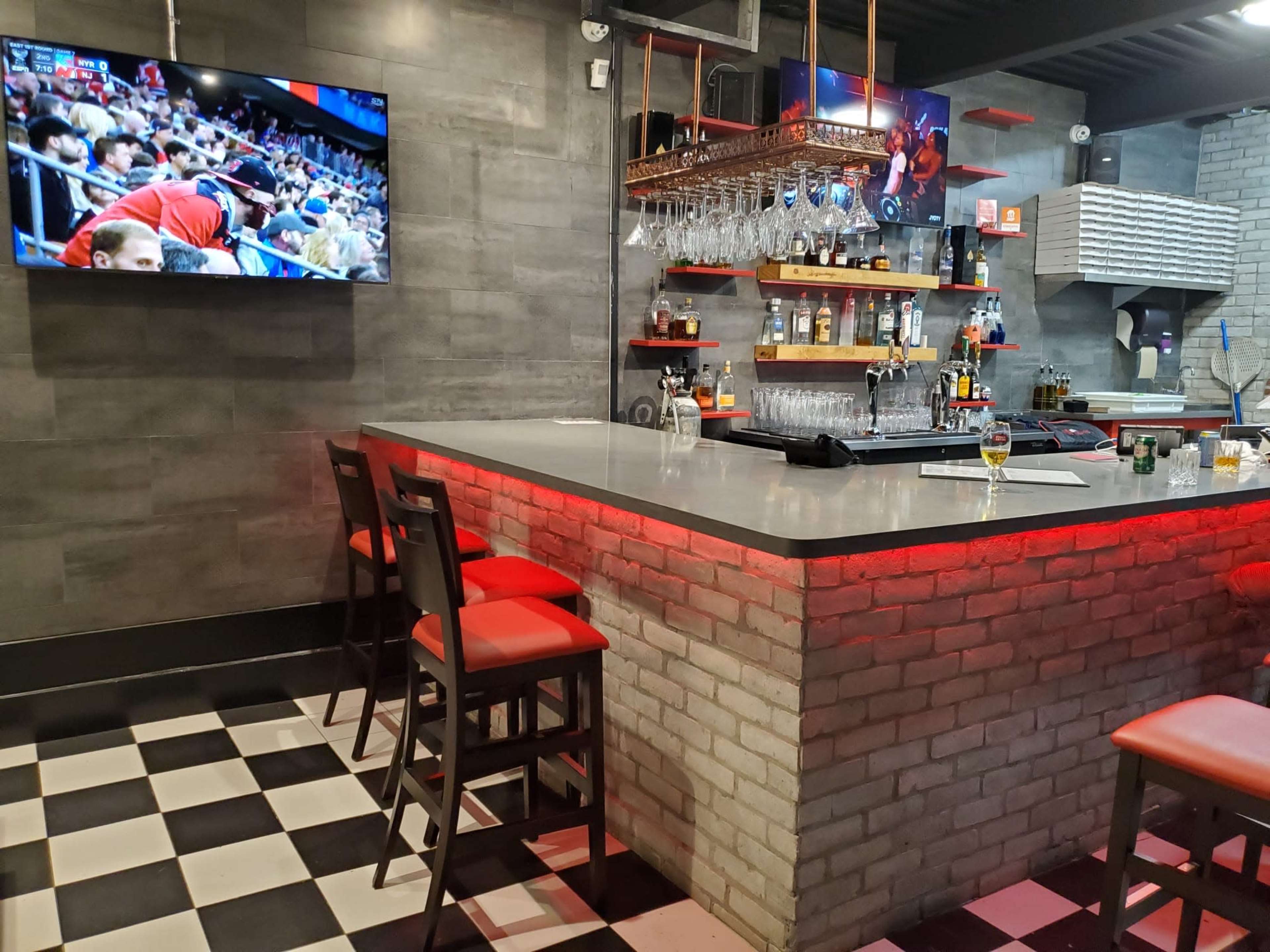 A bar area with a concrete wall, a television displaying a sports event, bottles on shelves, and red-lit seating.
