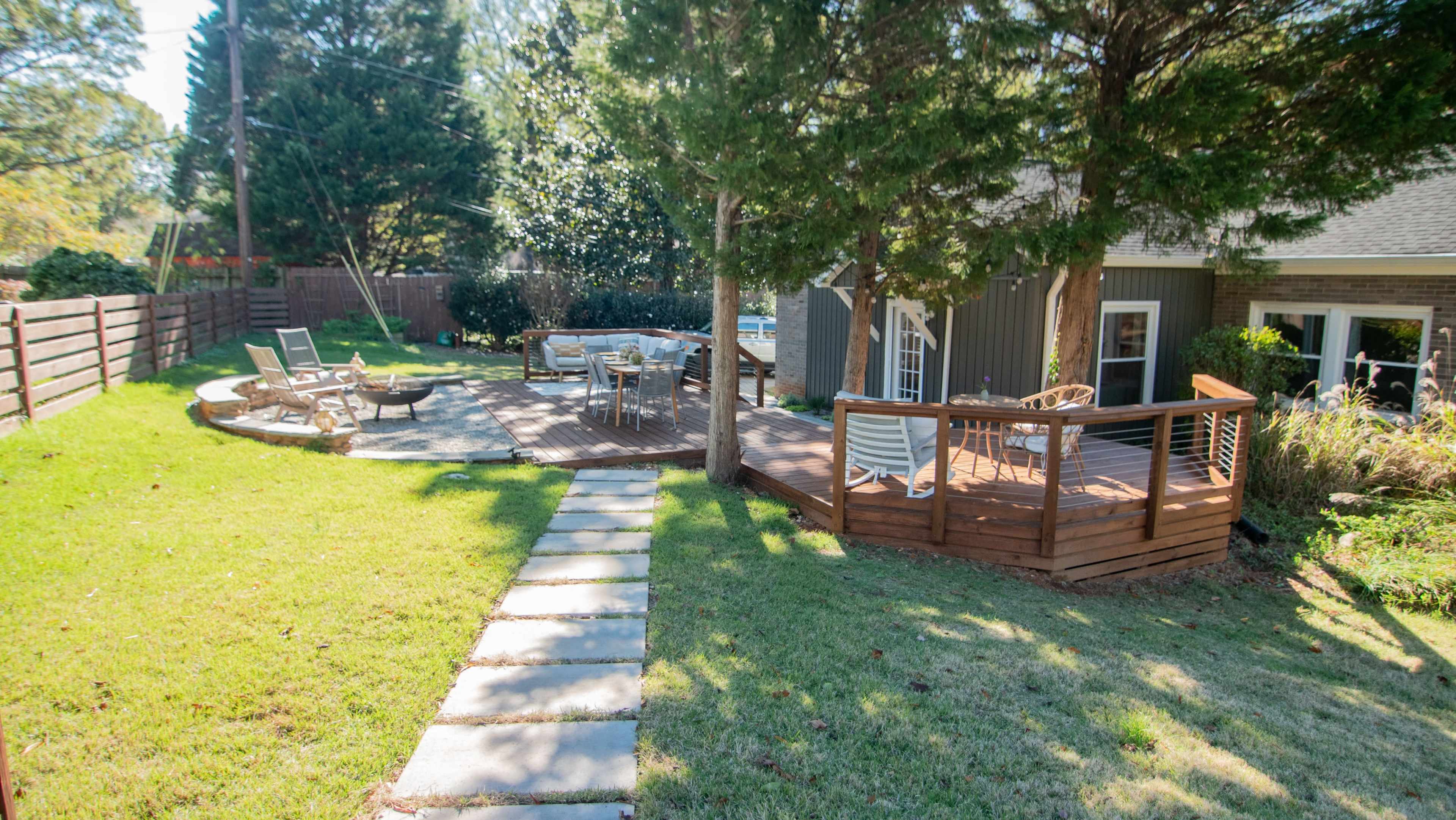 A backyard featuring a wooden deck with outdoor furniture, a stone pathway, and a circular seating area surrounded by grass and trees.