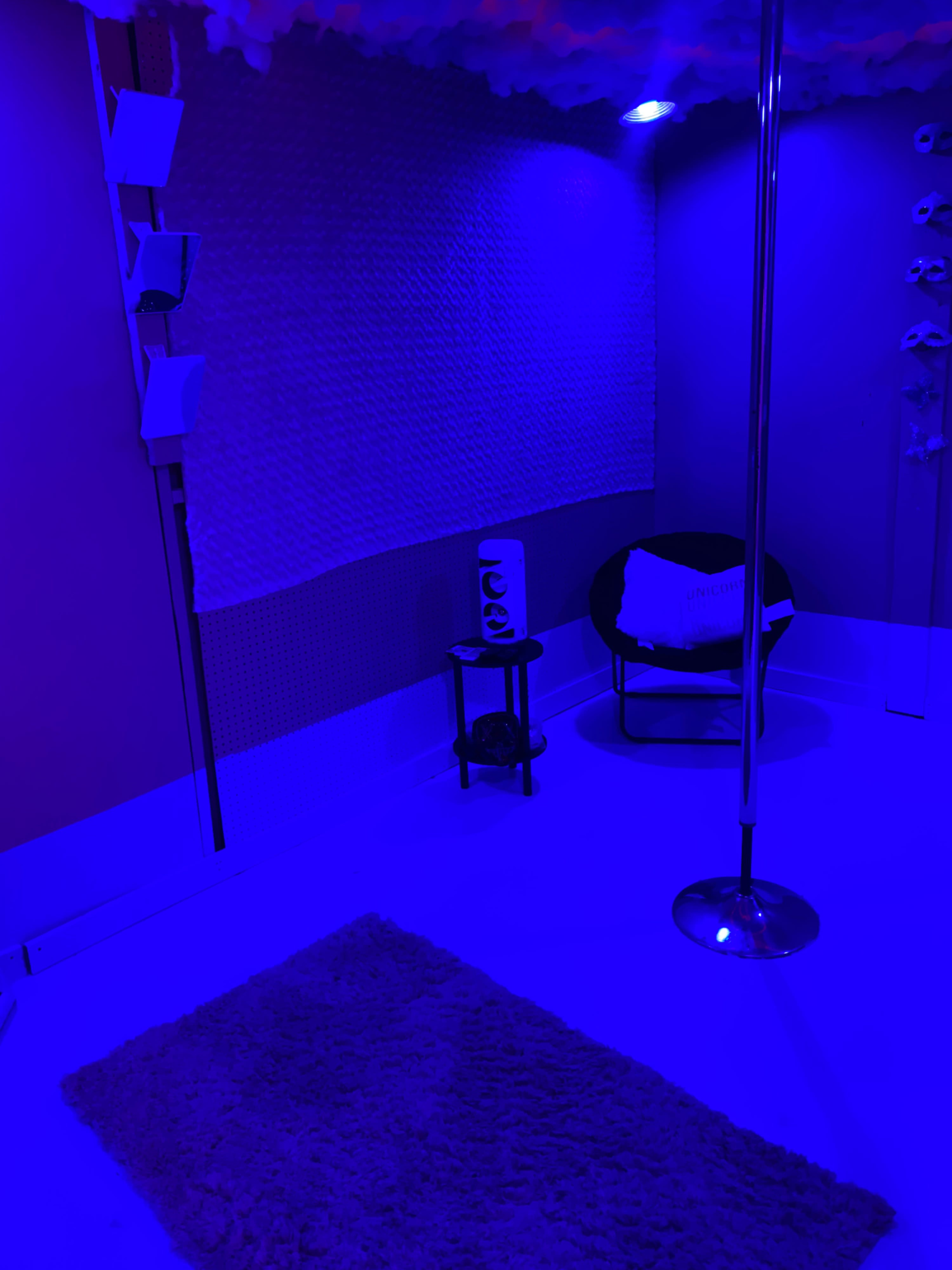 The image shows a dimly lit room with blue lighting, featuring a pole, a rug on the floor, a small table, and a chair.