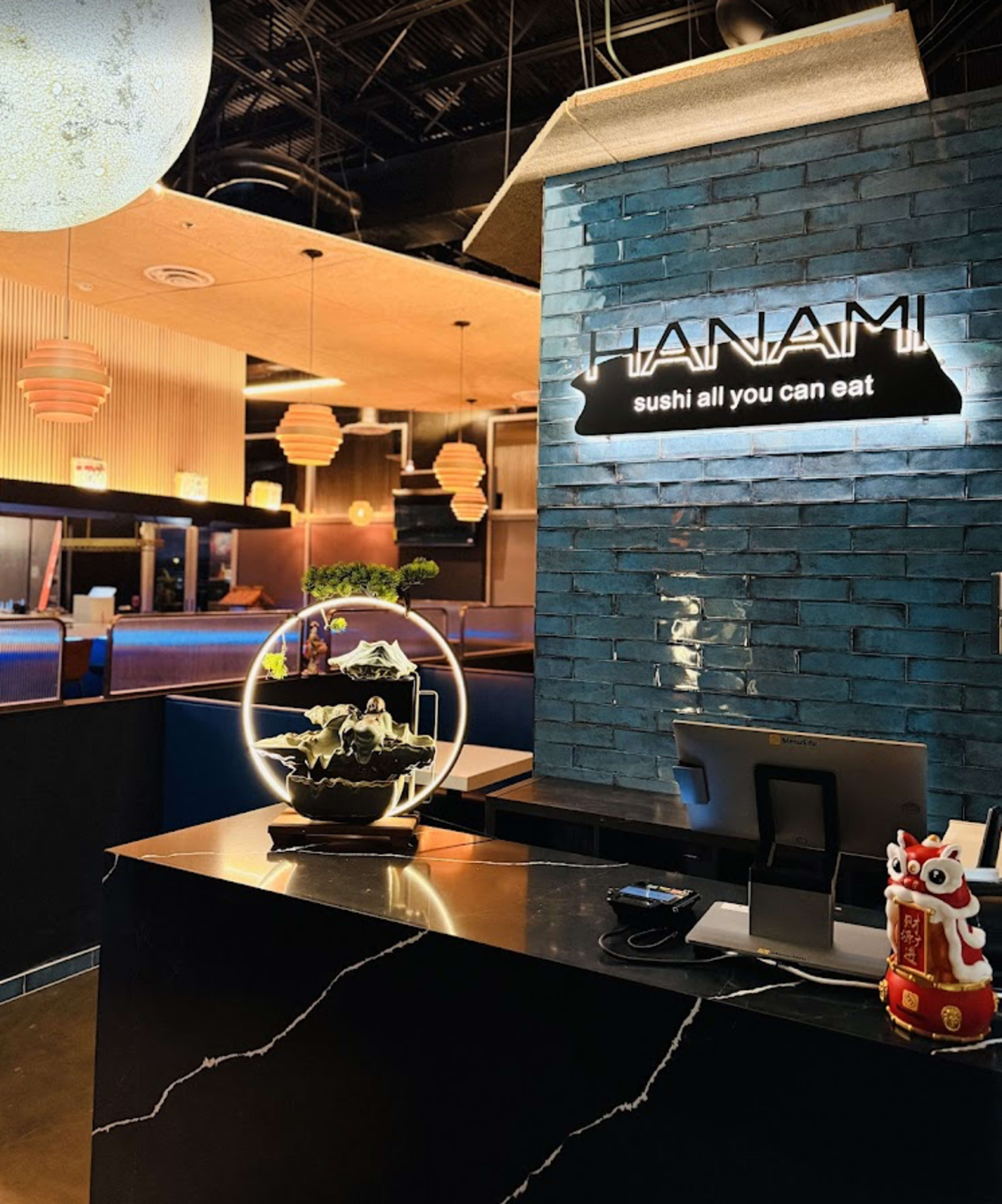 The image shows the interior of a sushi restaurant named "HANAMI," featuring a modern design with a reception area that includes decorative elements like a large spherical display and a detailed aquatic sculpture.