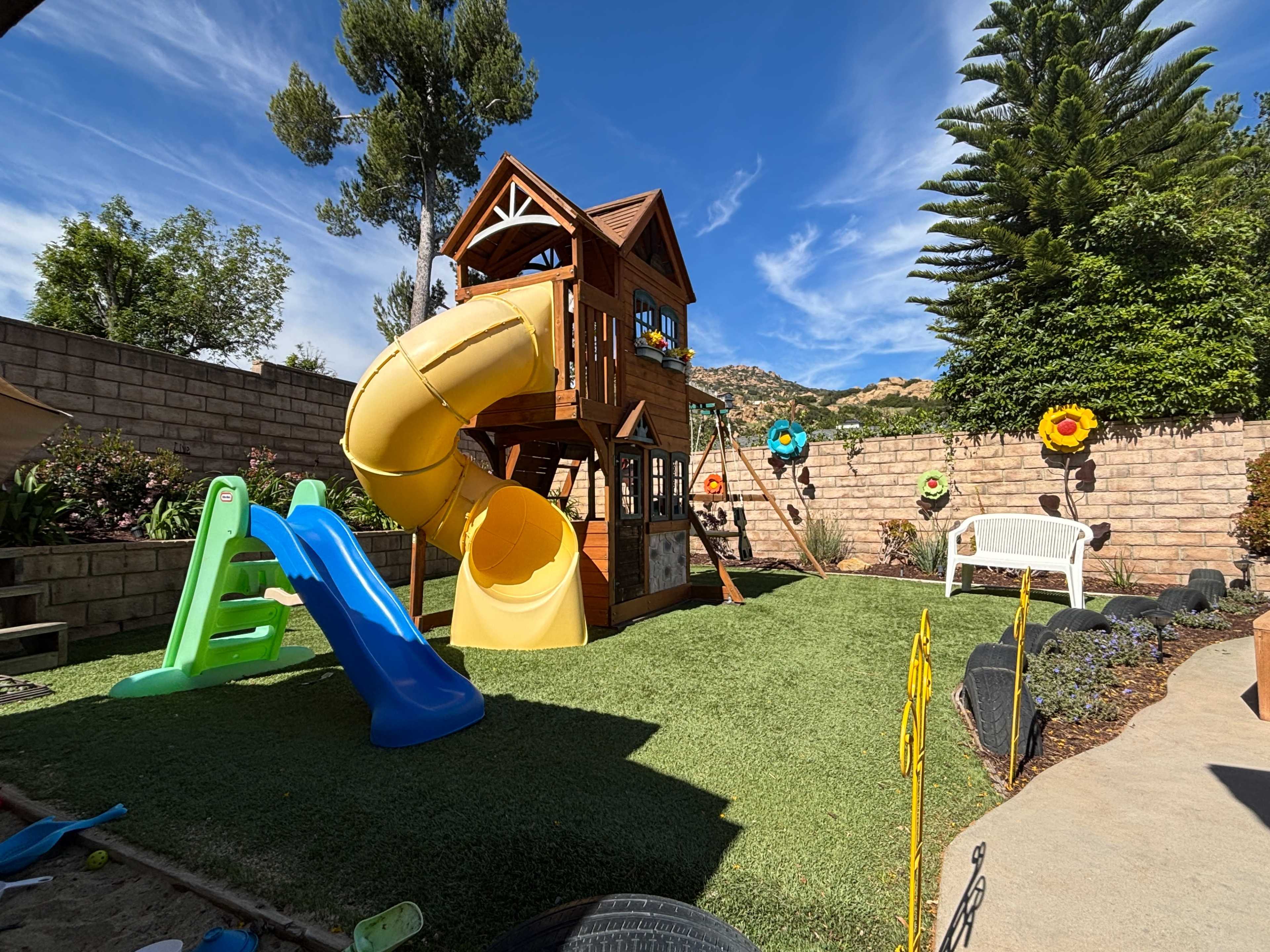The image shows a playground structure with a yellow slide and climbing features, surrounded by a grassy area and decorative flowers in a backyard setting.