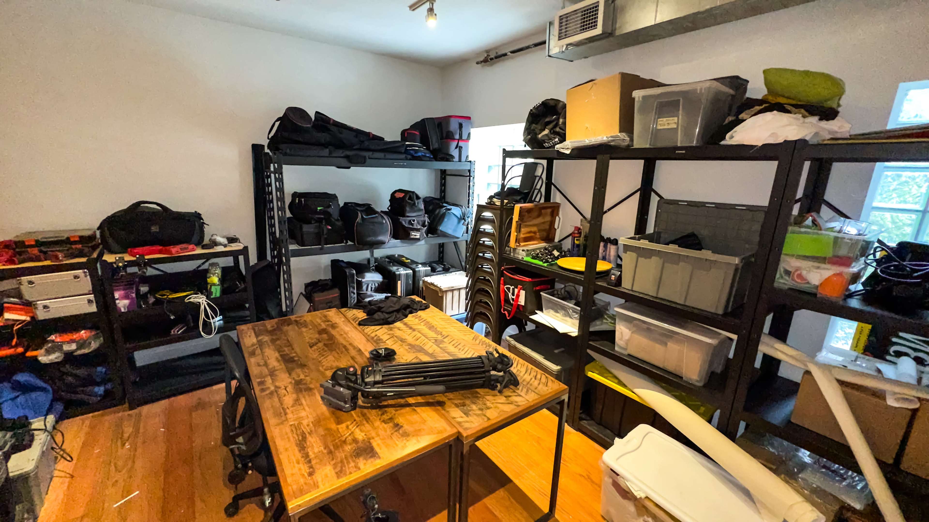 The image shows a cluttered room with shelves filled with various bags and equipment, and a wooden table in the center.
