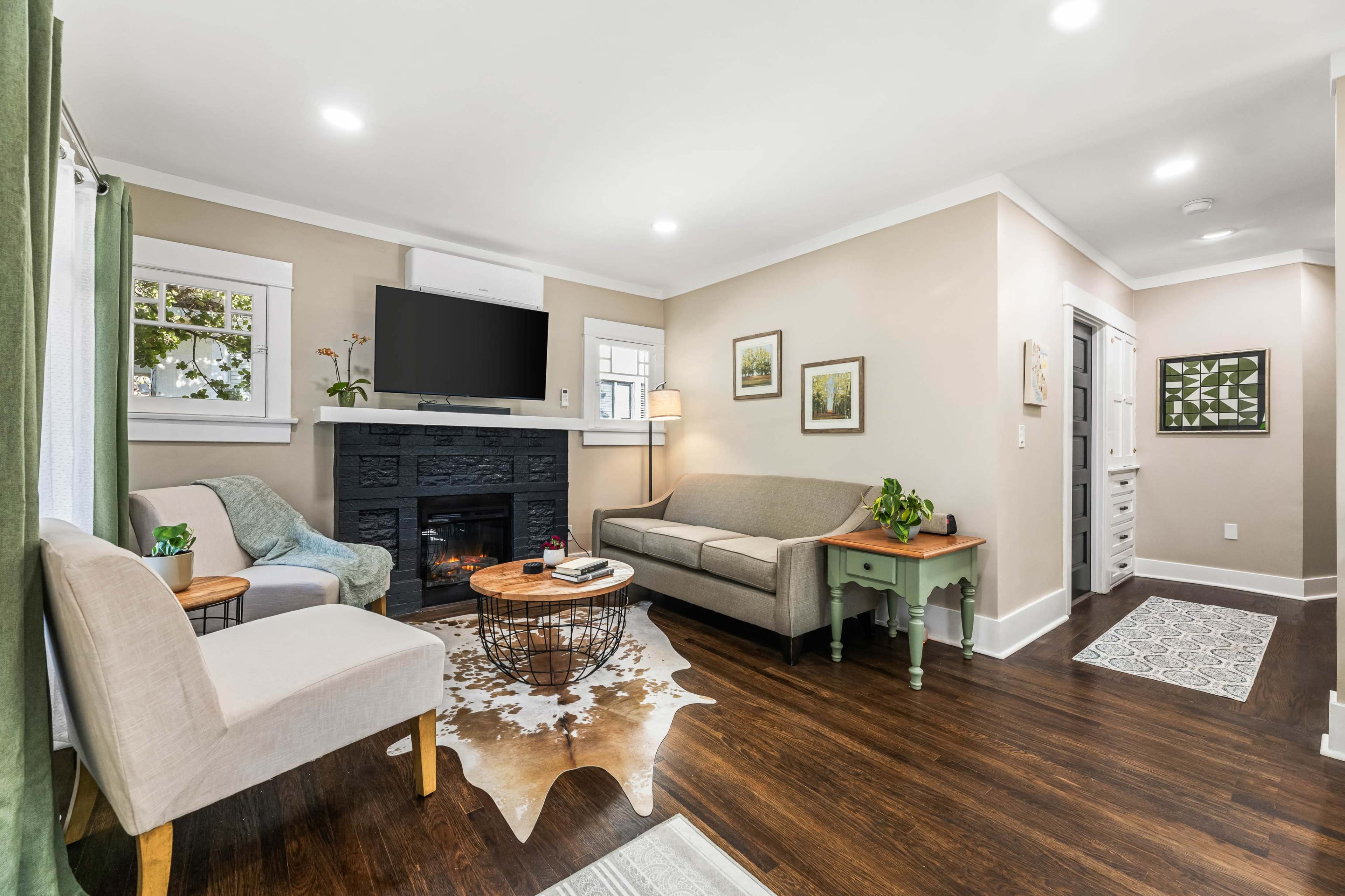 A cozy living room features a gray sofa, a light-colored armchair, a mounted TV above a black fireplace, and decorative artwork on the walls.