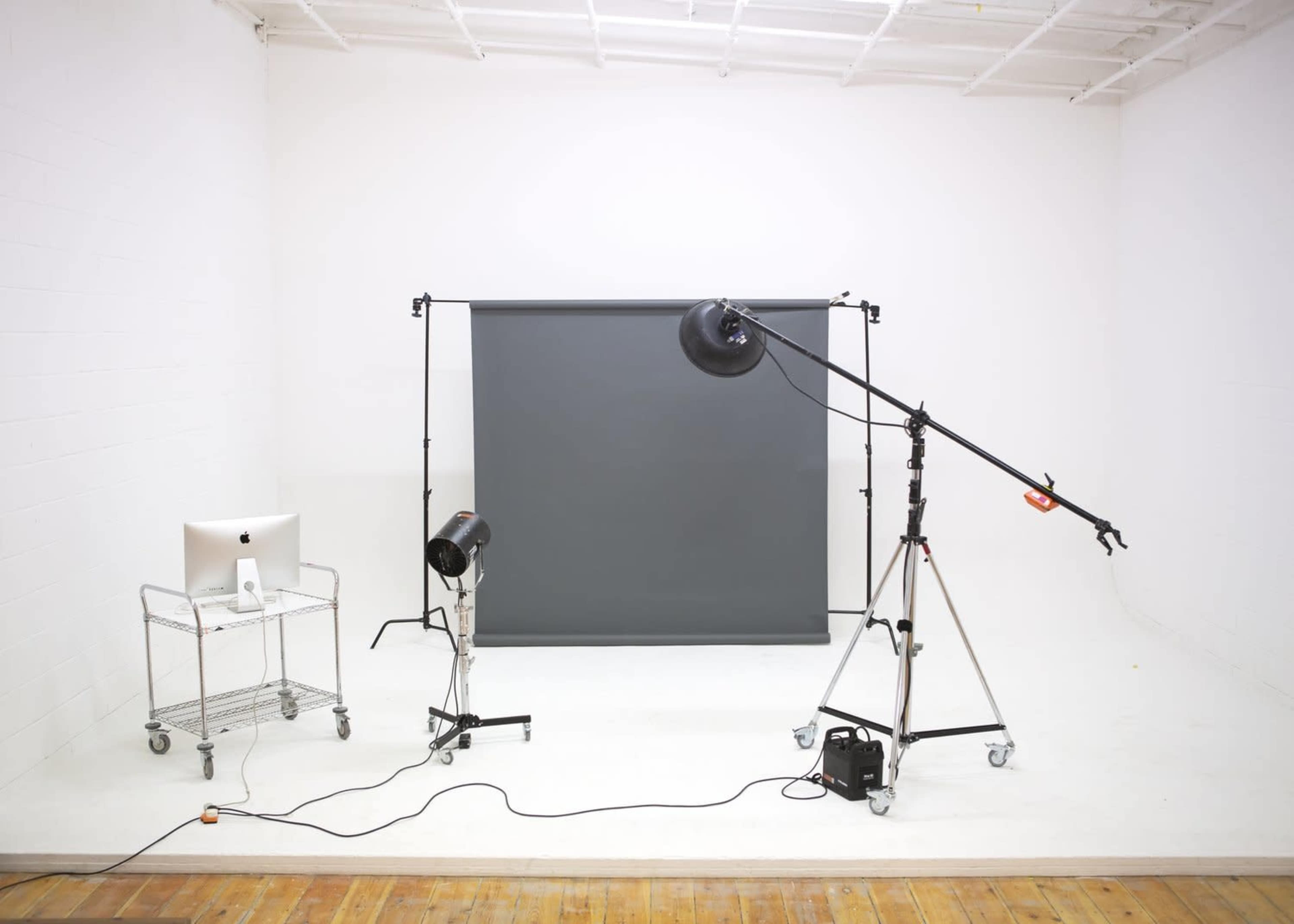 A photography studio is set up with a gray backdrop, lighting equipment on stands, and a computer on a cart.