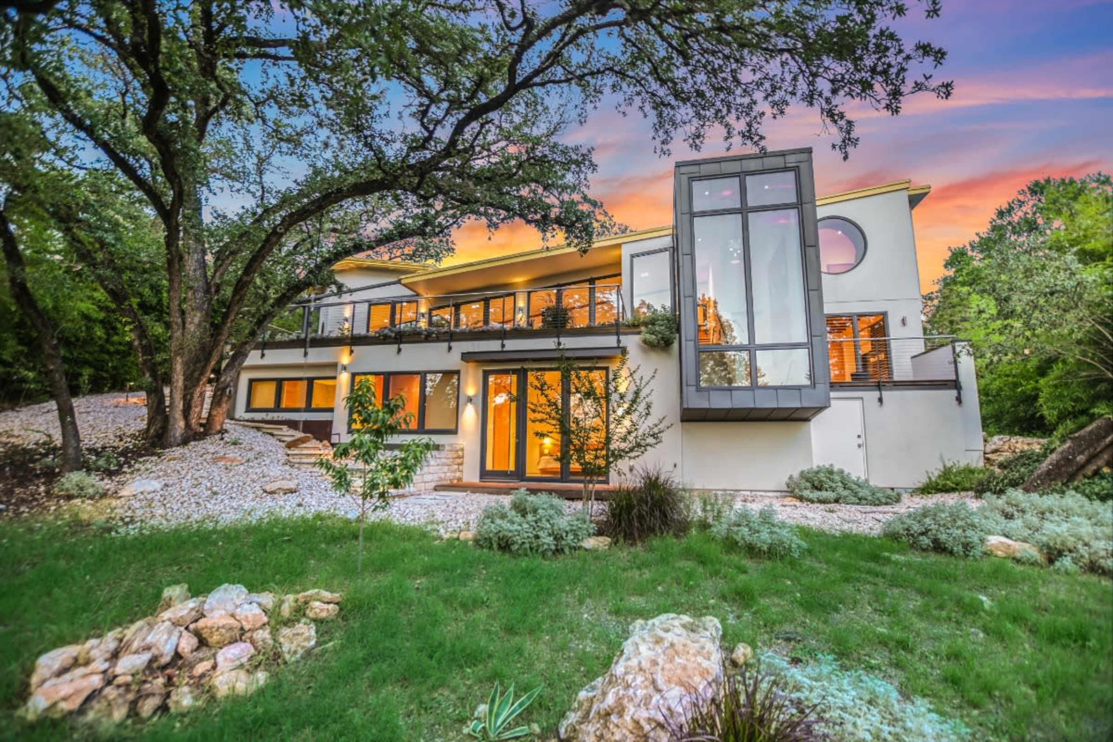 A modern two-story house with large windows and a landscaped yard is situated among trees and rocks, illuminated by a colorful sunset sky.