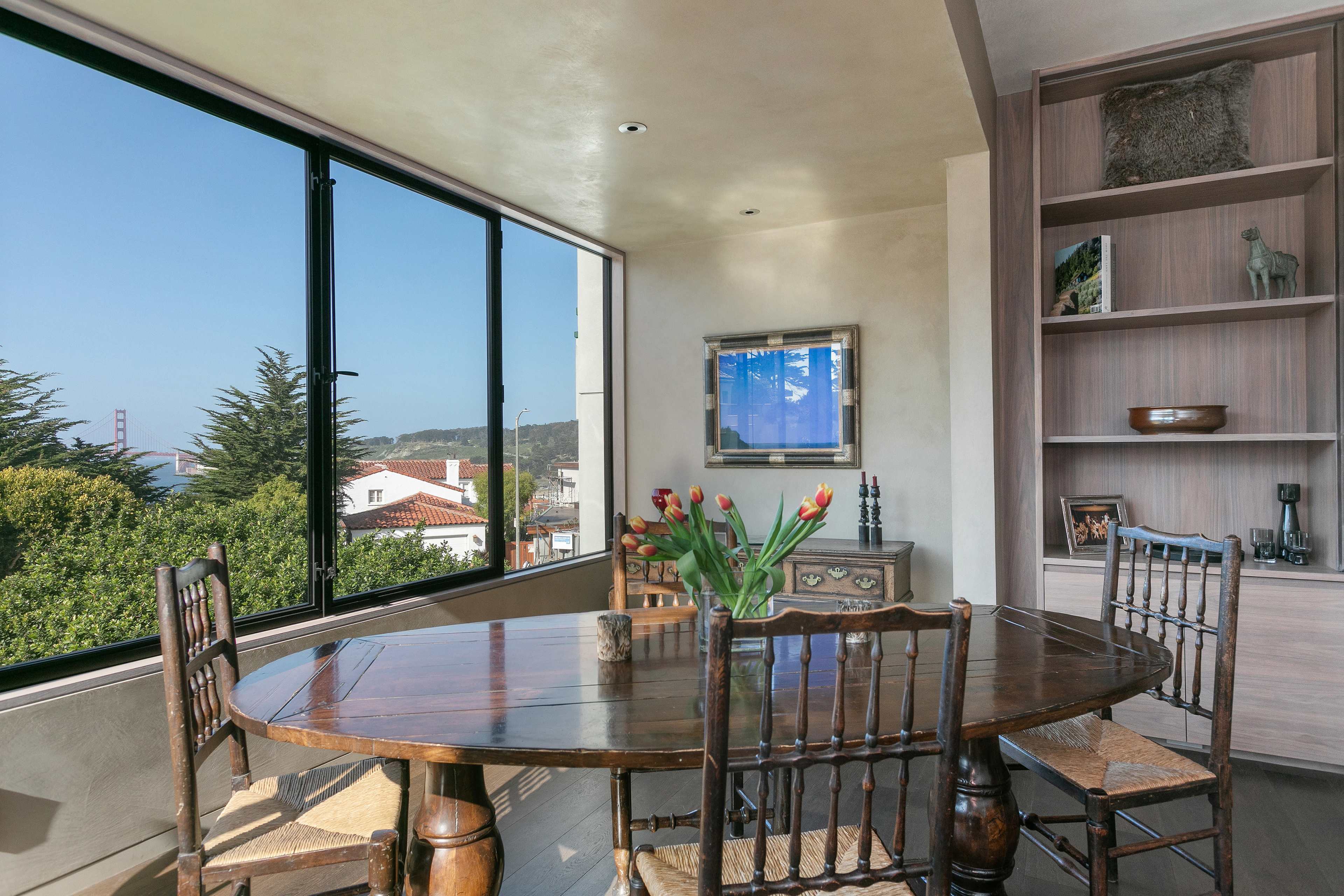 A wooden dining table with five chairs is positioned near large windows that offer a view of greenery and a distant bridge.