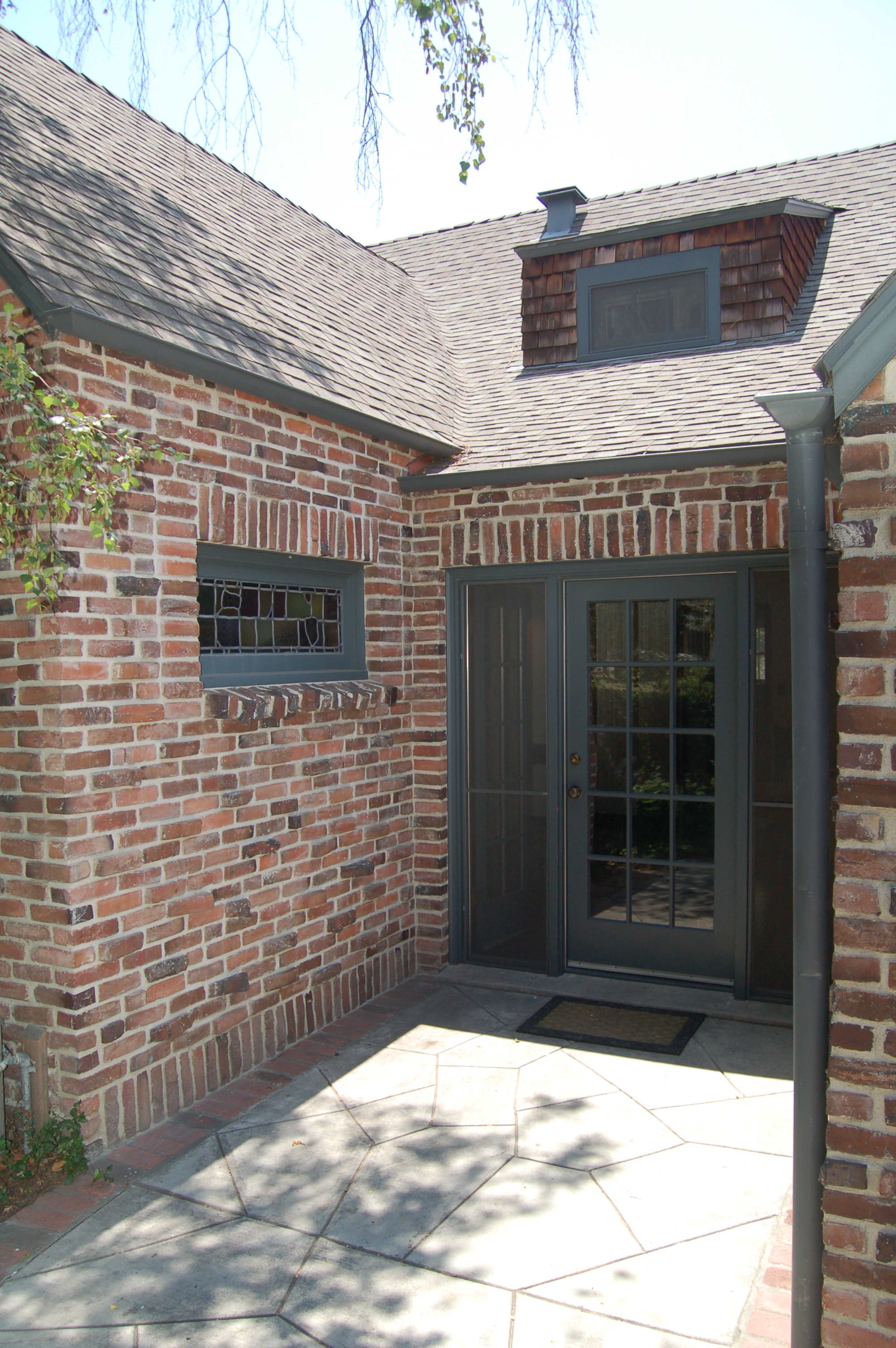 The image shows a brick wall exterior of a house with a dark-framed glass door and a small window, set against a stone pathway.
