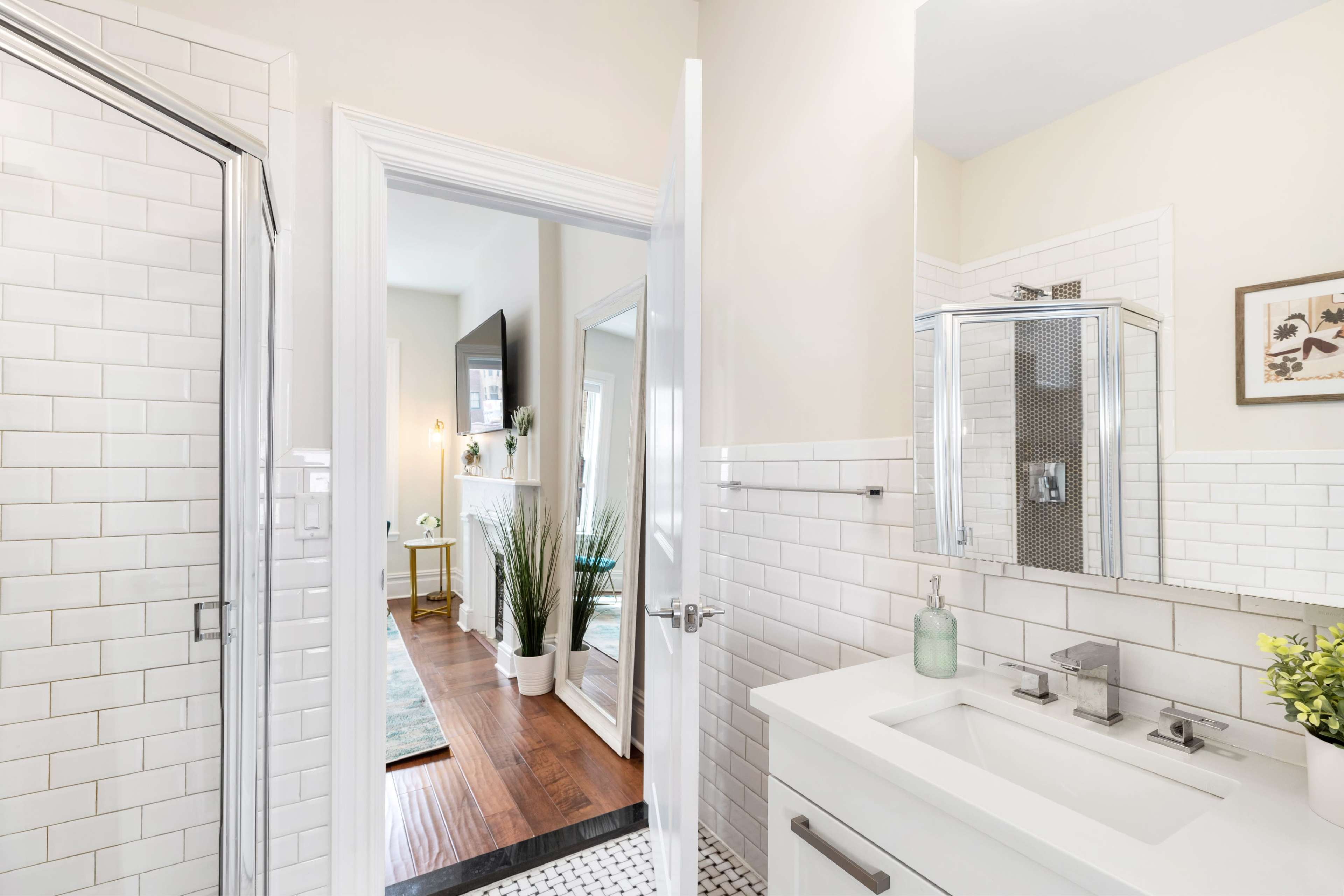 A modern bathroom with white subway tile walls, a glass shower, and a doorway leading to a well-lit adjoining room.