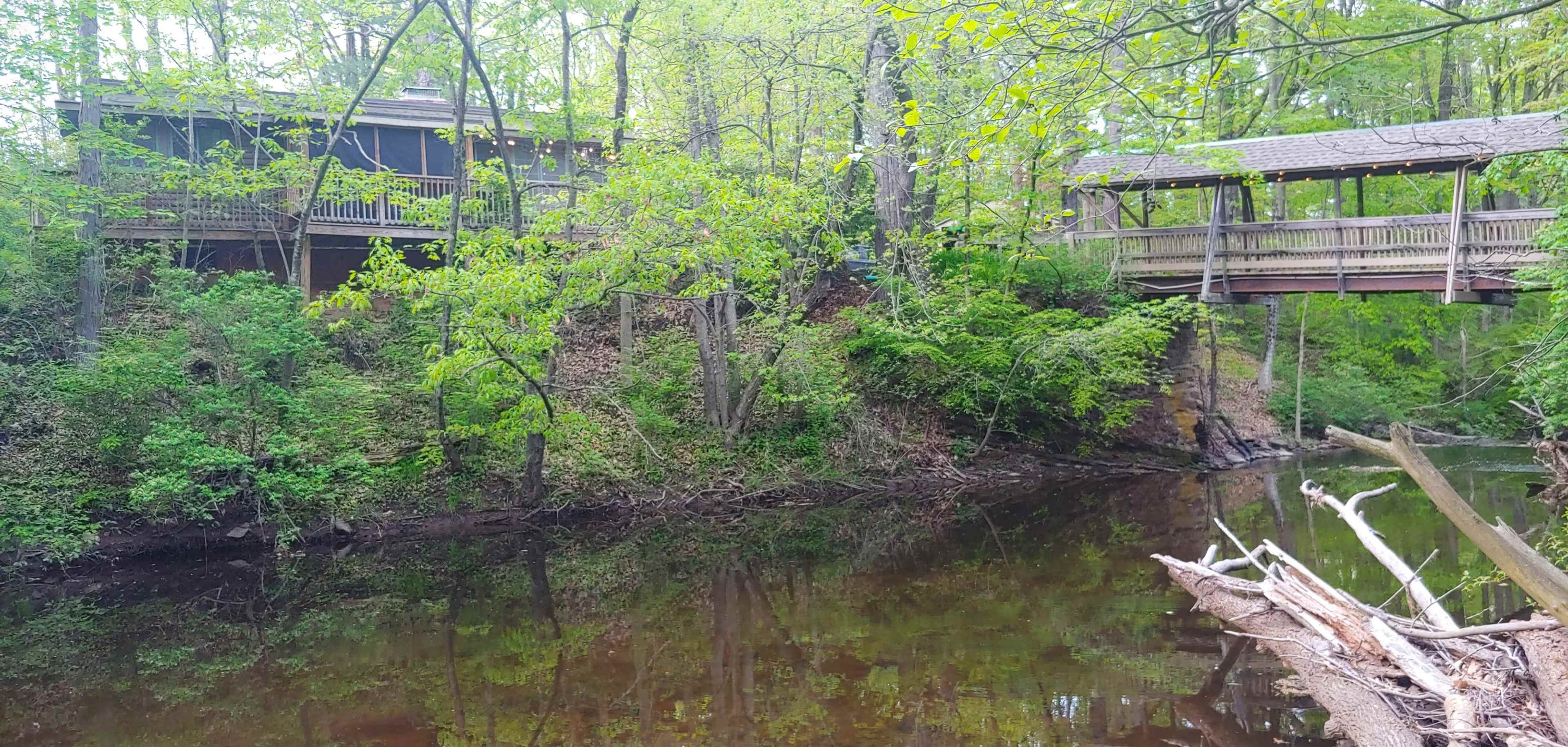 A wooden bridge connects a house on the shore to a platform above a calm river, surrounded by lush greenery.