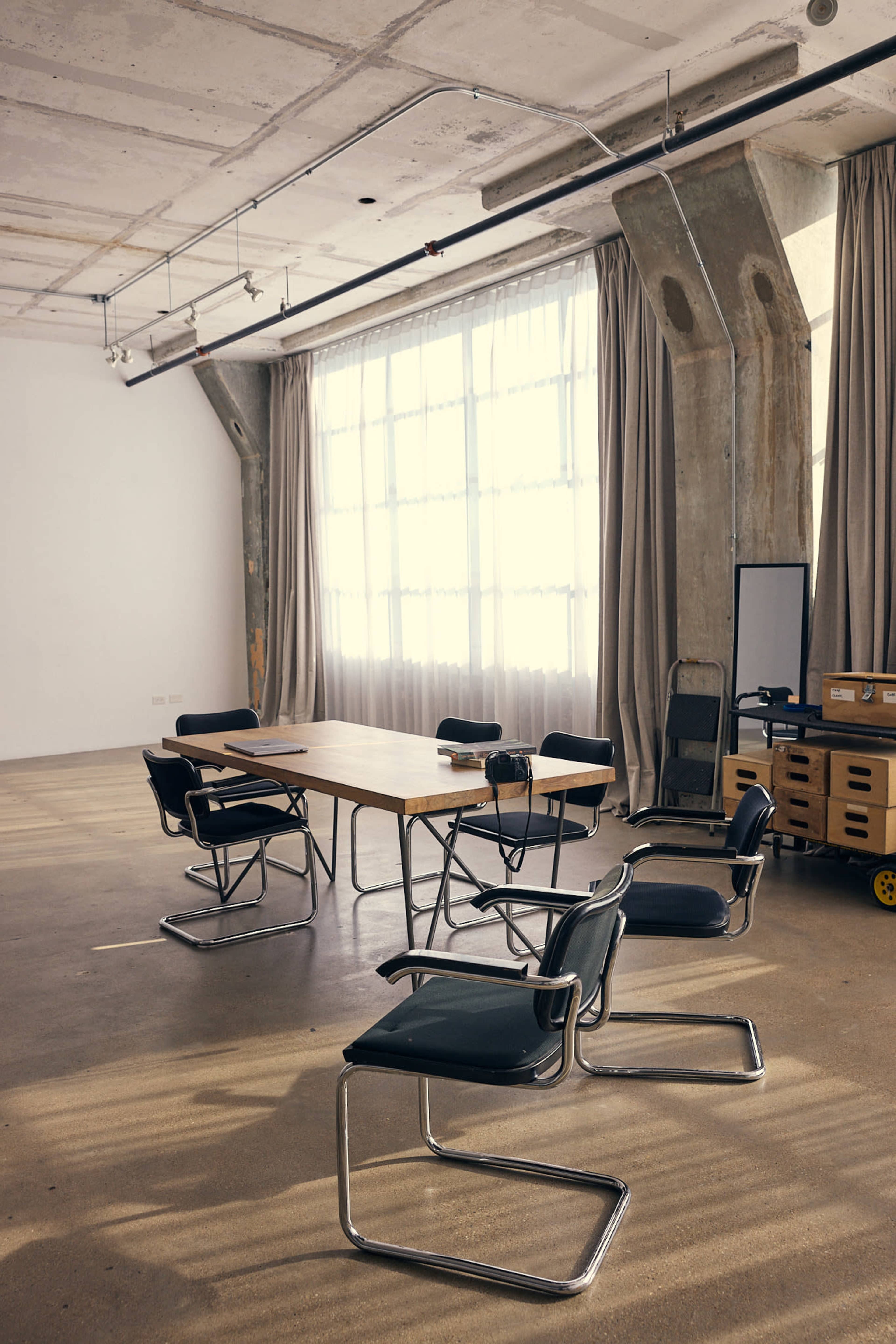 A modern office space features a wooden table surrounded by black chairs, with large windows and concrete walls.