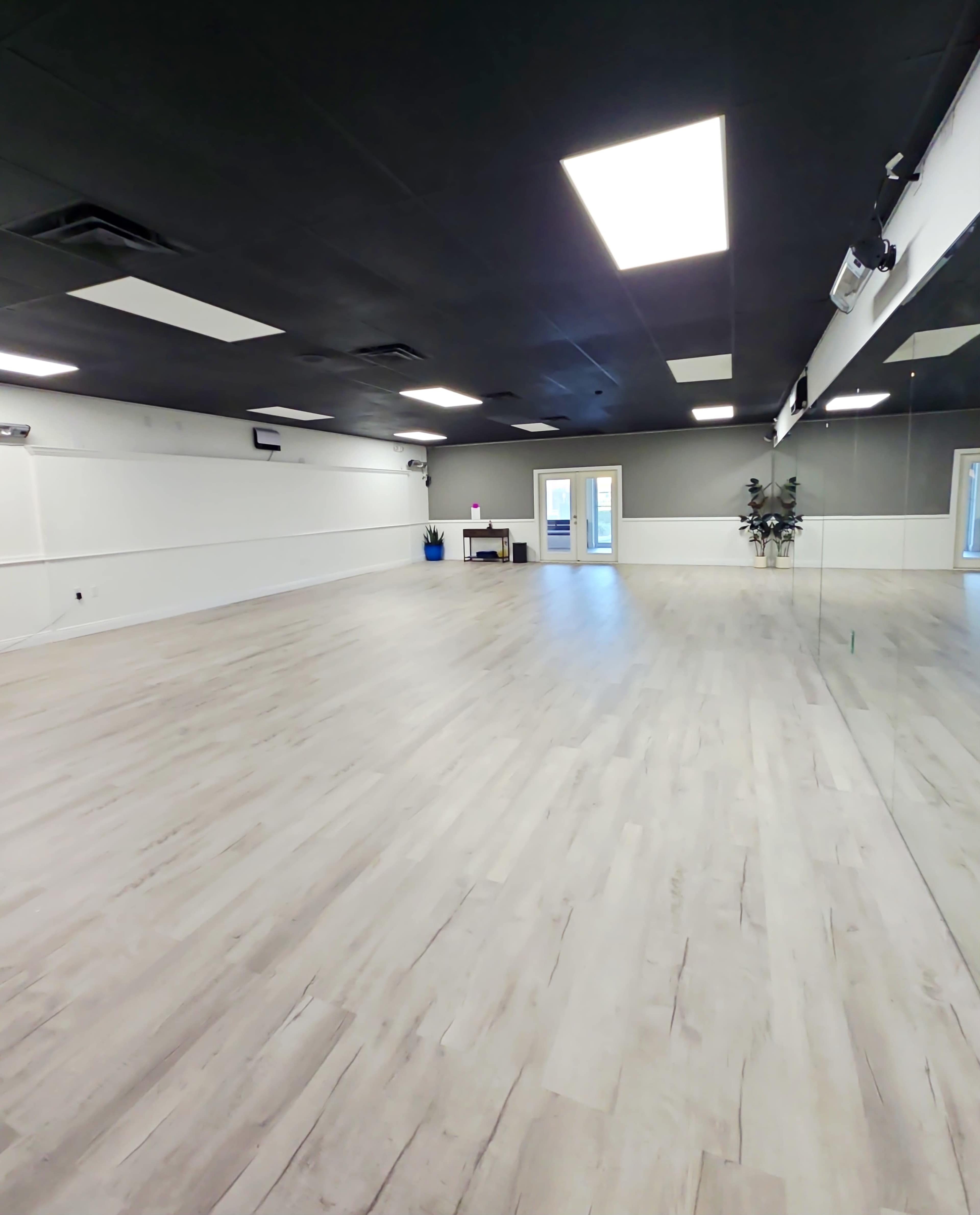 The image shows a spacious, empty studio with wooden flooring, large mirrors along one wall, and bright overhead lights.