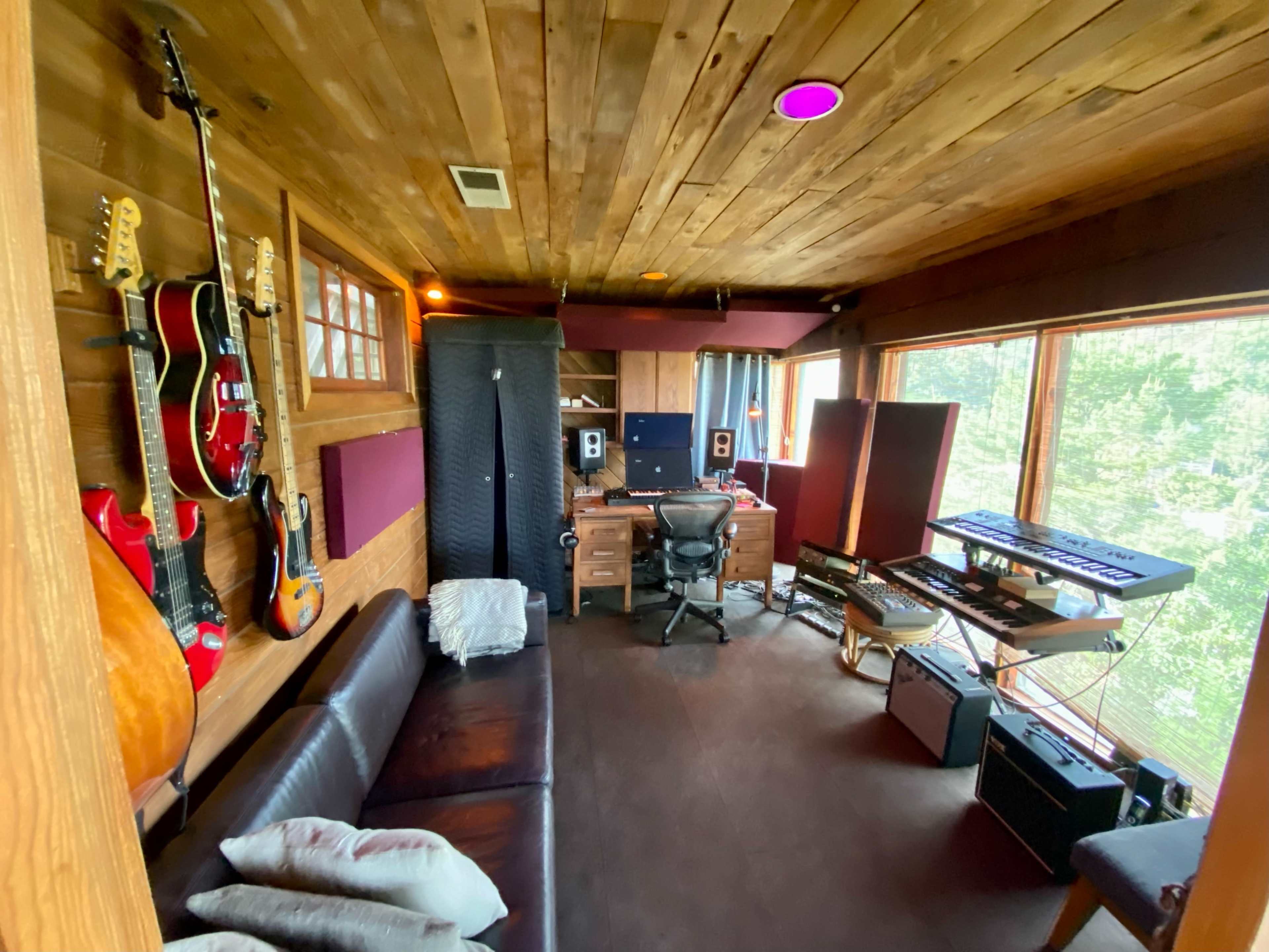 The image shows a music recording studio with wooden walls, a desk setup with audio equipment, guitars hanging on the wall, and large windows offering a view of nature.