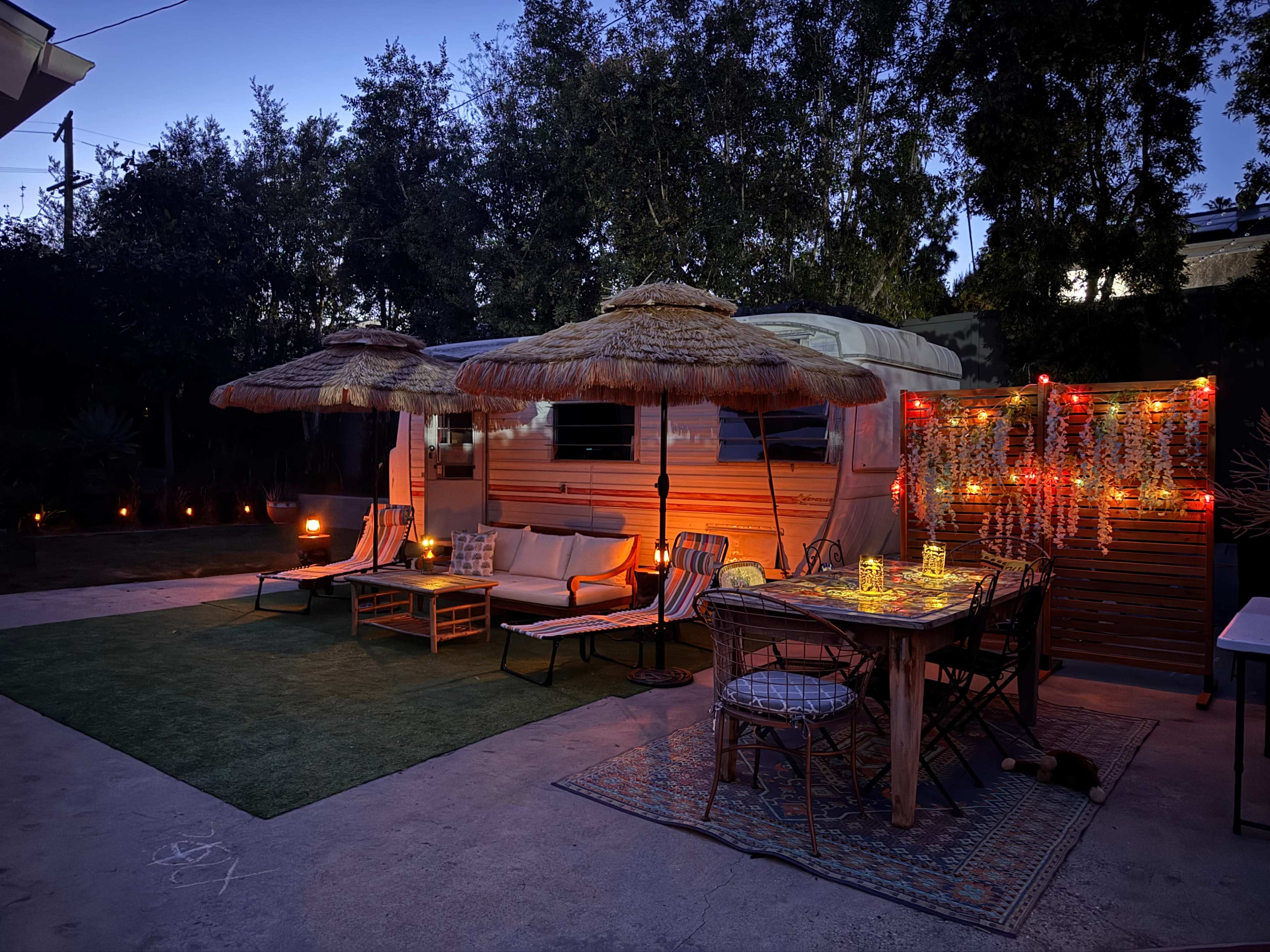 A cozy outdoor space features a retro trailer surrounded by patio furniture, two thatched umbrellas, and soft lighting, creating an inviting atmosphere.