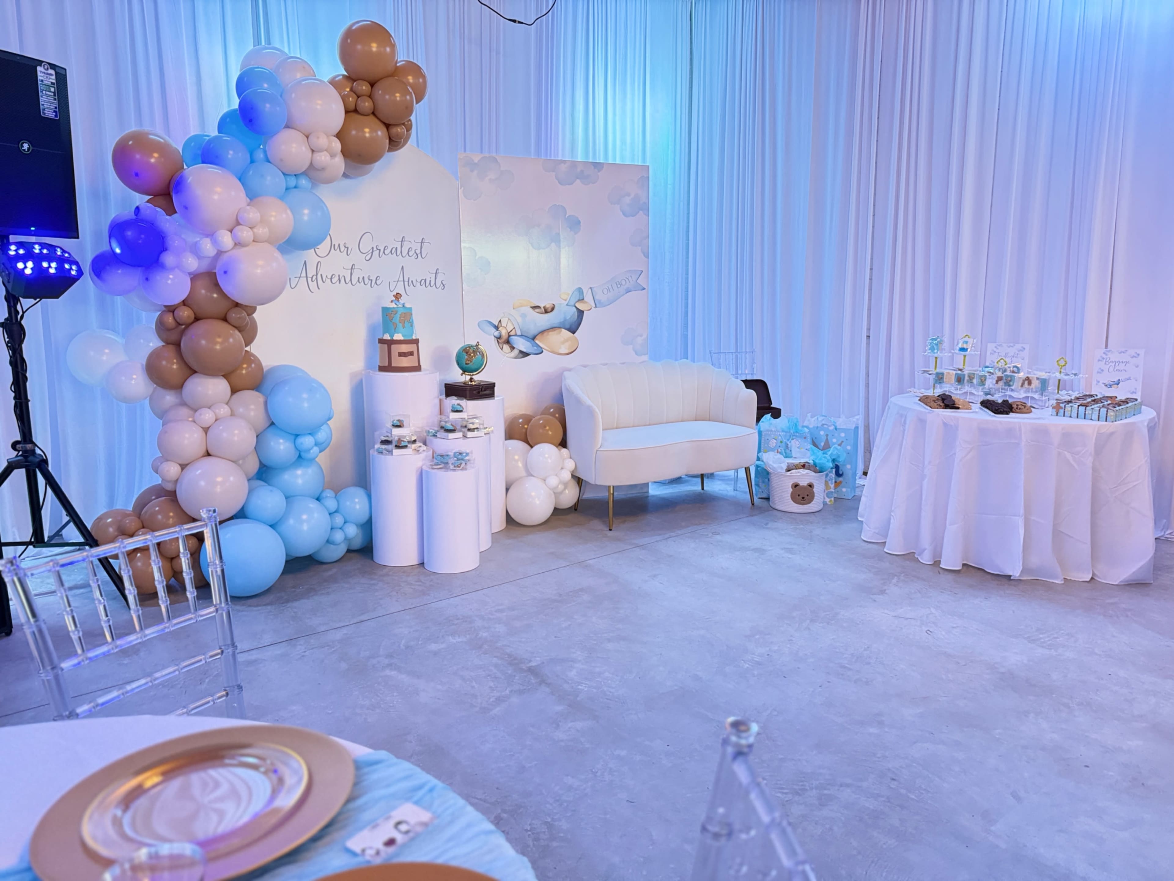 The image shows a decorated event space featuring a balloon arch in blue and brown, a white couch, a dessert table with treats, and a backdrop with a cloud and balloon design.