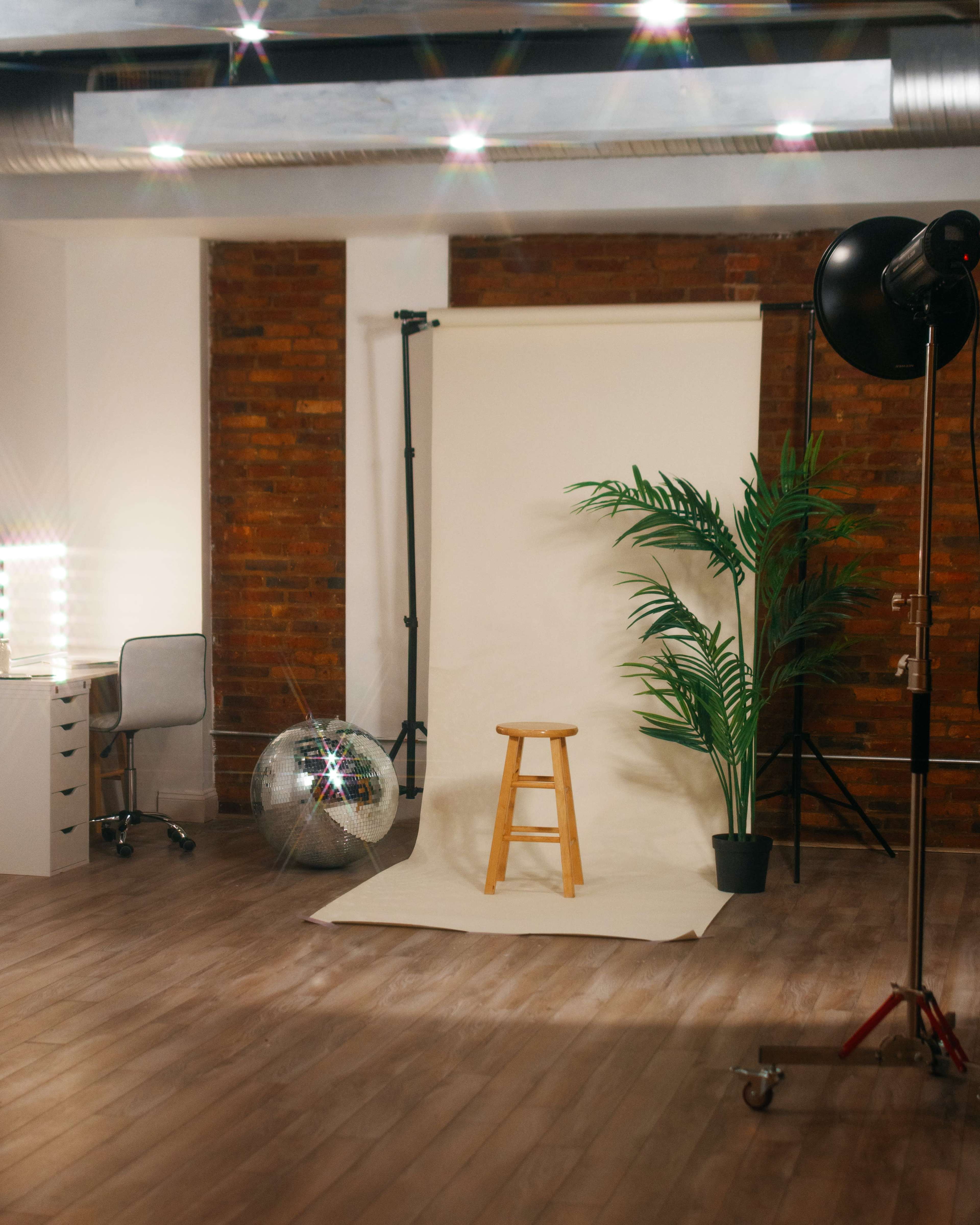 A photography studio with a wooden stool in front of a backdrop, a plant in a pot, a disco ball, and a makeup station with lights.