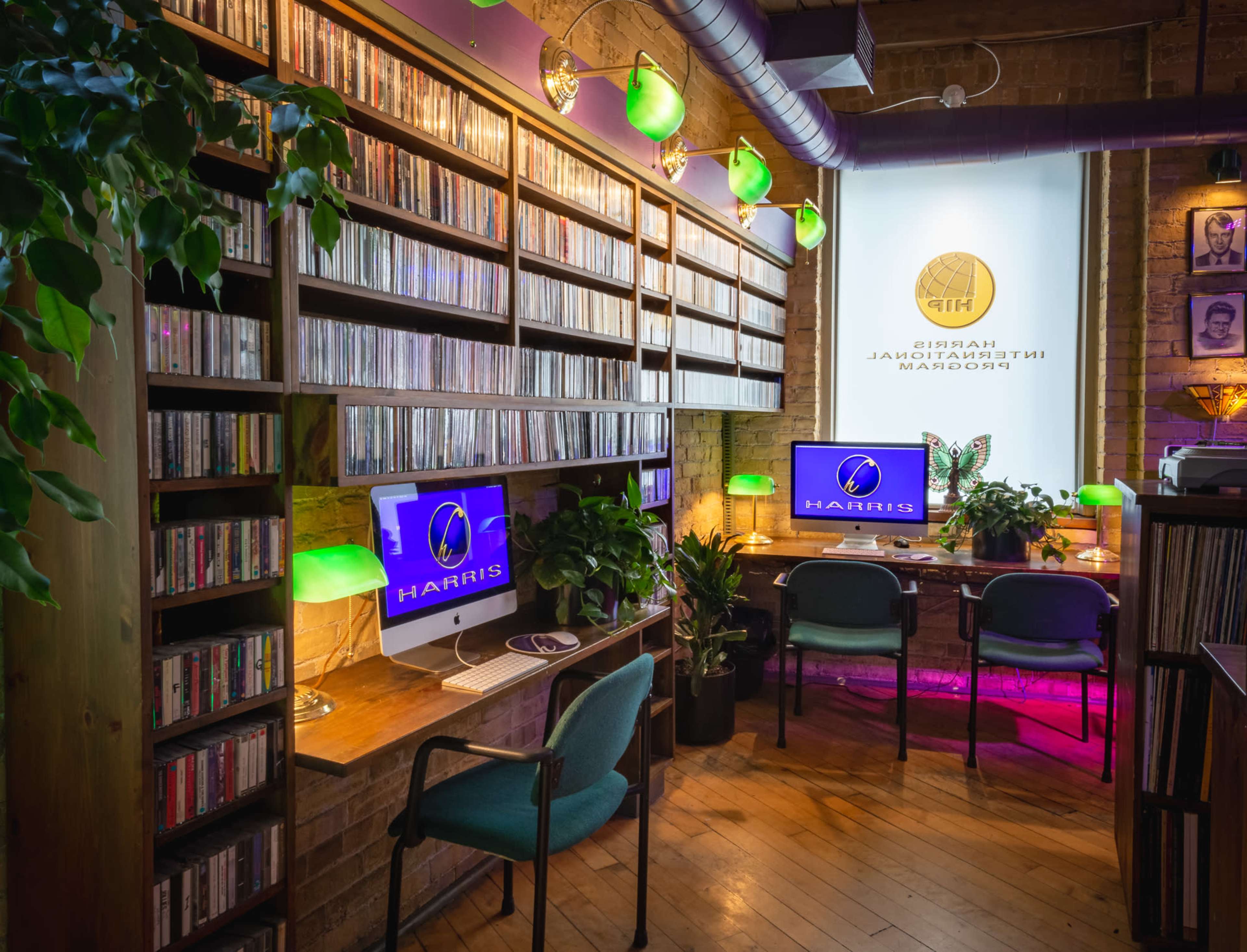 A cozy workspace features shelves filled with CDs, two computers with monitors displaying the word "HAIRIS," and green lamps providing ambient lighting.