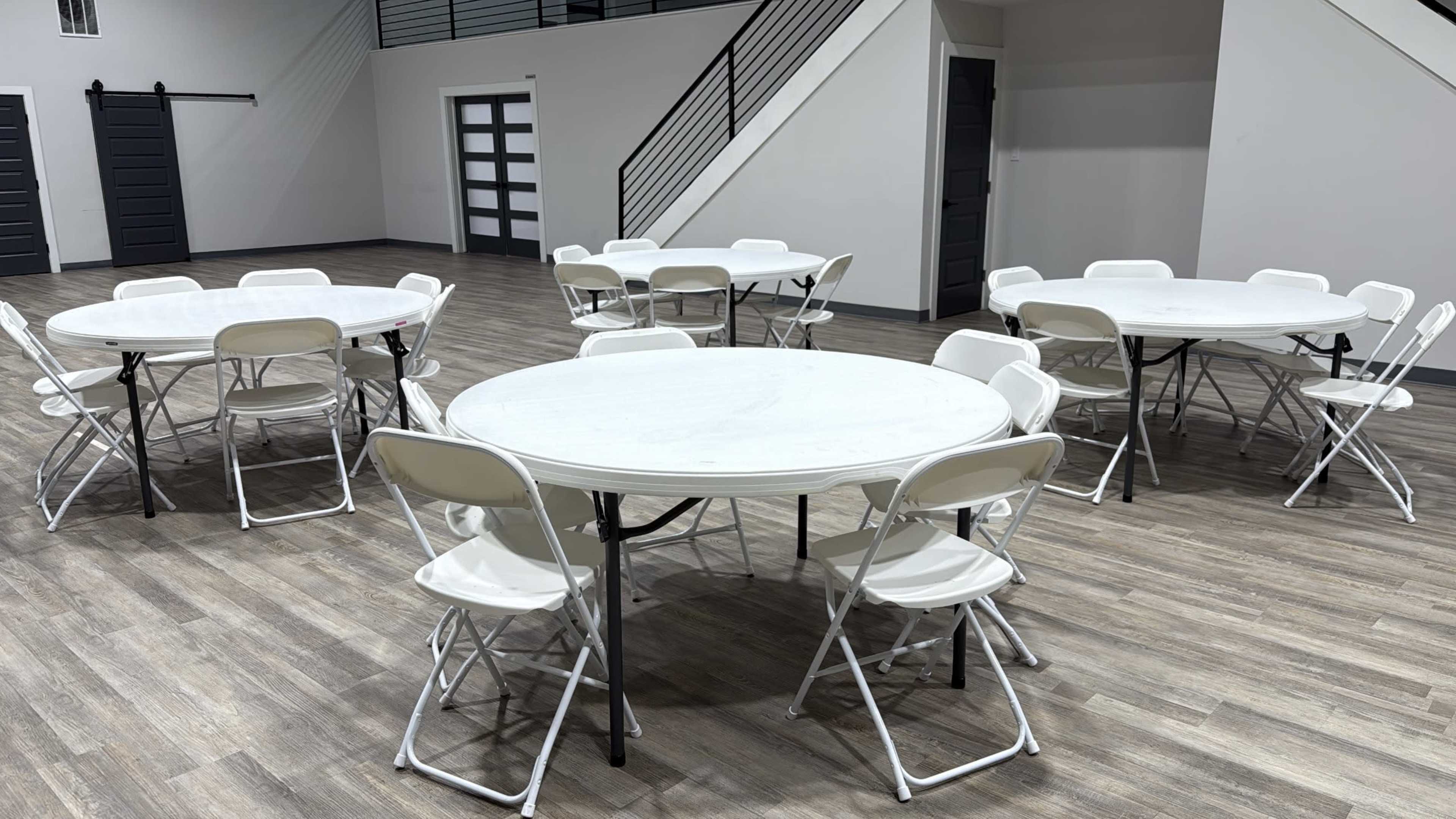 The image shows a spacious room with several round tables surrounded by white folding chairs on a wooden floor.