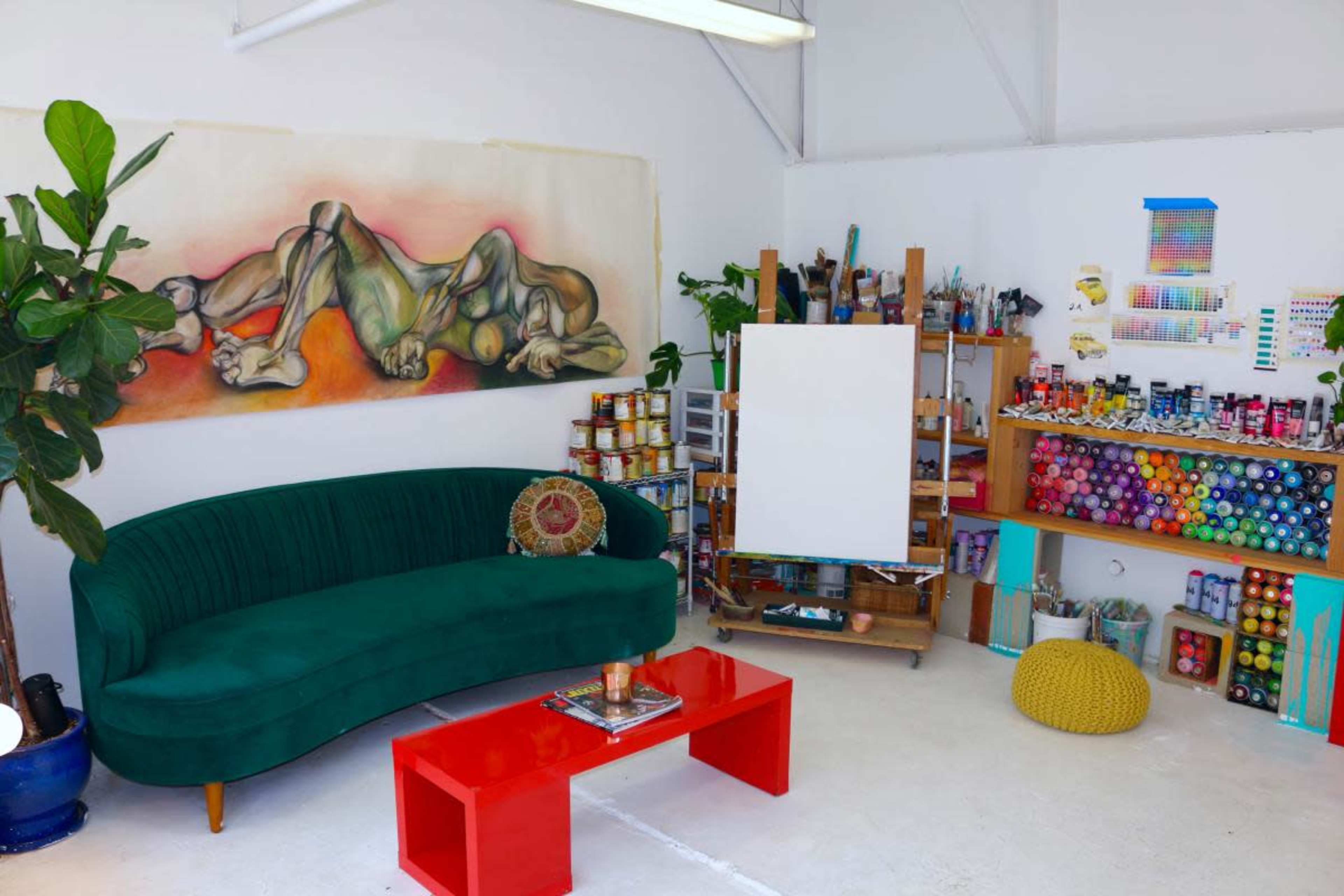 A bright art studio featuring a green velvet couch, a red coffee table, an empty canvas, and shelves stocked with various paints and art supplies.