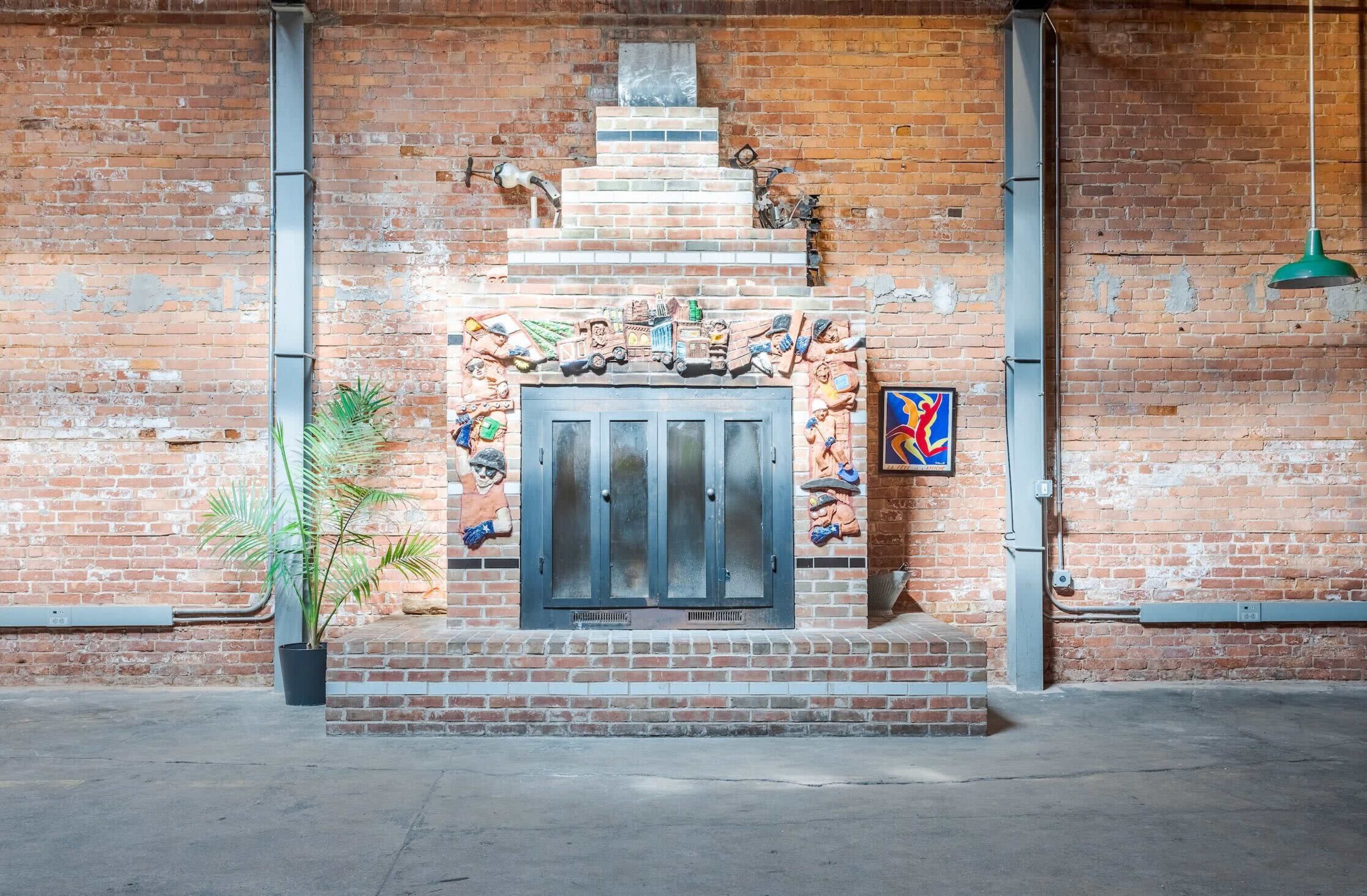 A brick wall with a decorated fireplace bordered by sculptures, accompanied by a potted plant and a colorful artwork on the side.