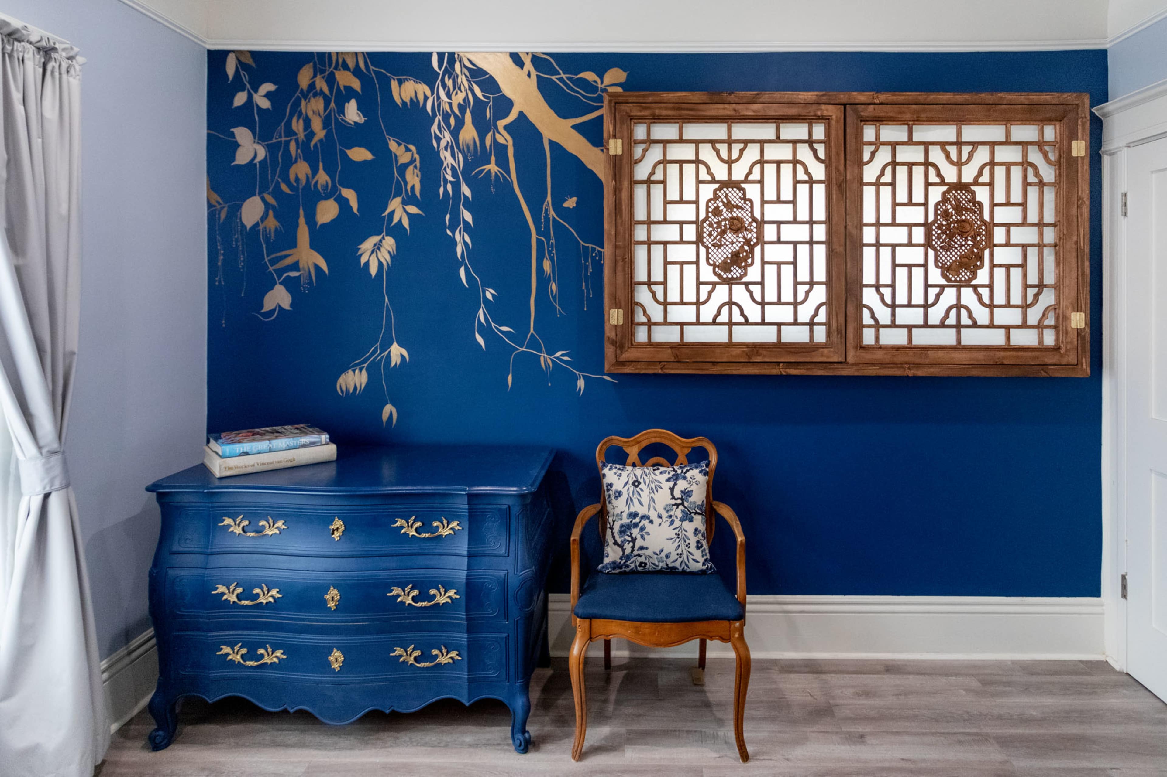 A blue accent wall adorned with gold botanical illustrations features an ornate wooden window frame above a blue dresser and a wooden chair with a decorative cushion.