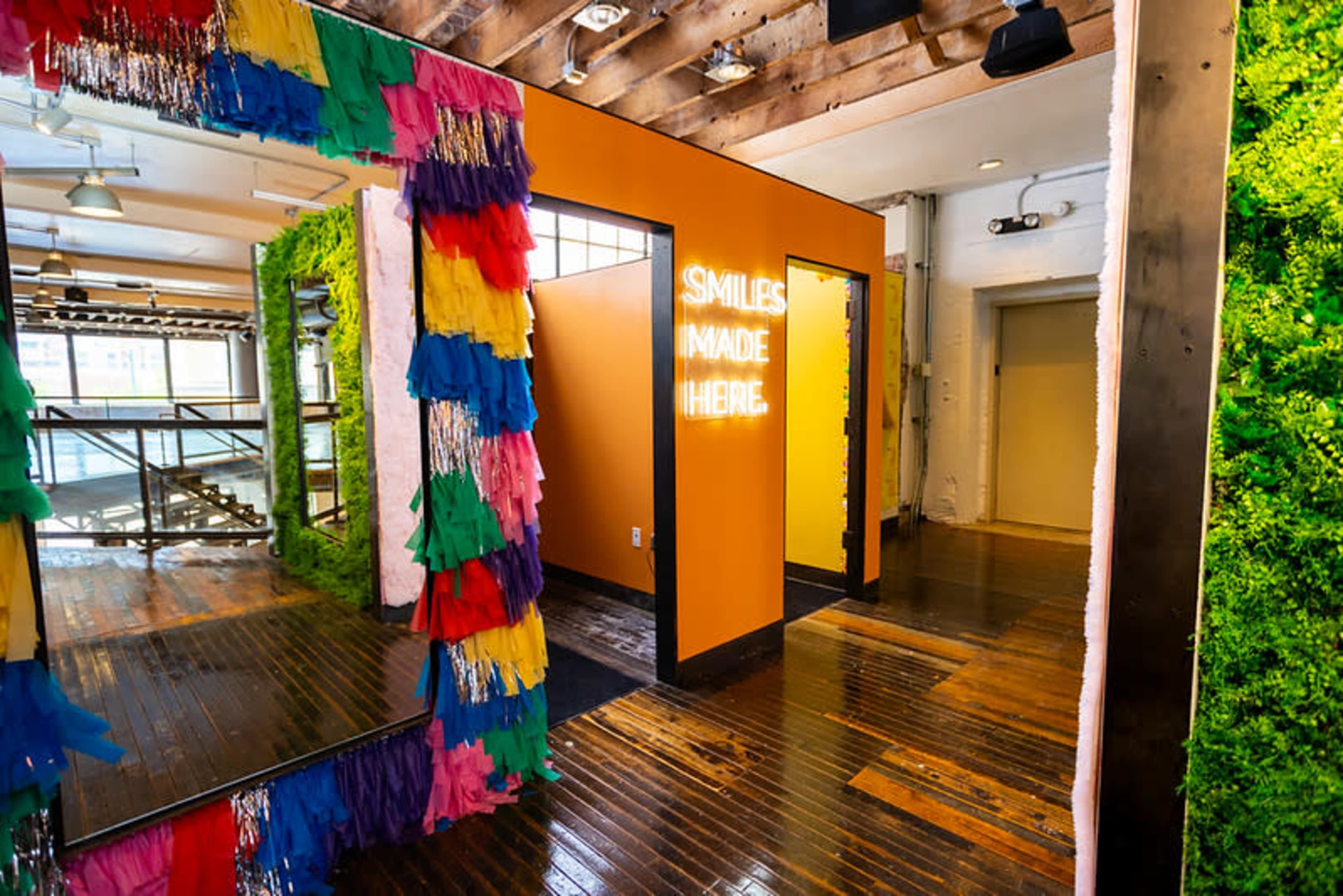 The image shows a colorful interior space featuring mirrors with fringe decorations, illuminated signage that reads "SMILES MADE HERE," and a vibrant moss wall.