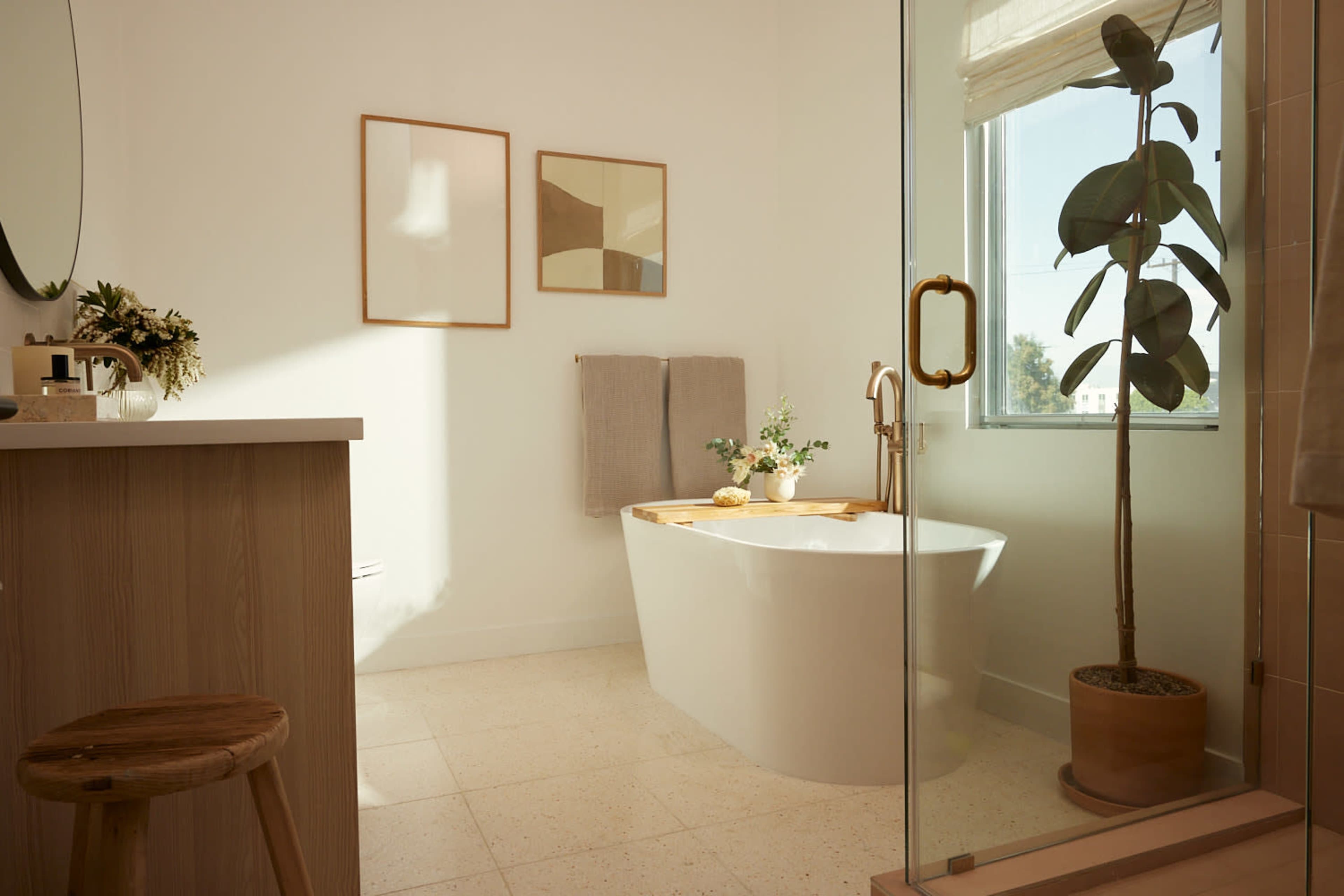 A bright bathroom featuring a freestanding tub, a wooden counter with a stool, two framed artworks, and a potted plant near a window.