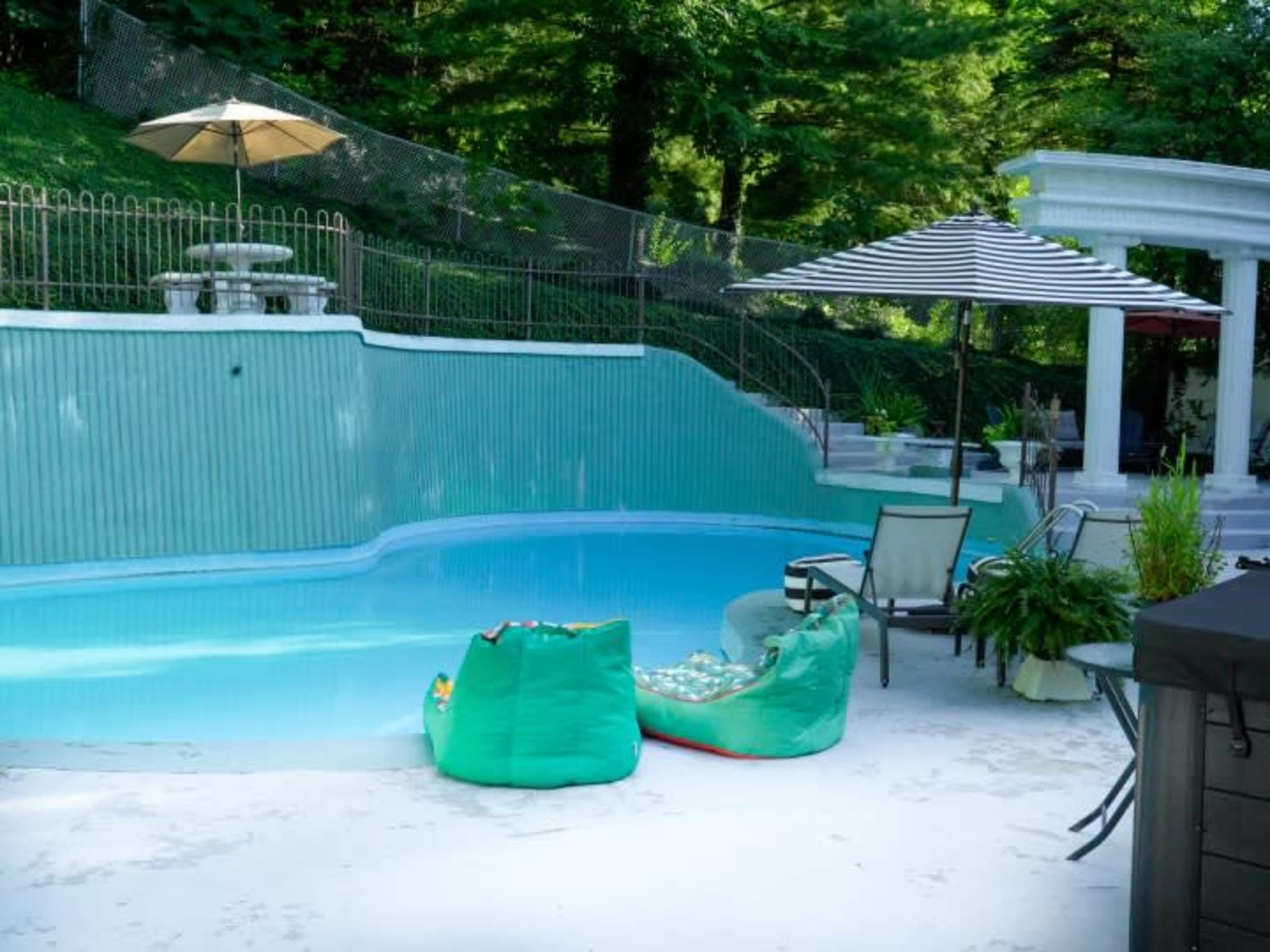 A pool area surrounded by green trees, featuring a light blue pool, lounge chairs, and umbrellas.