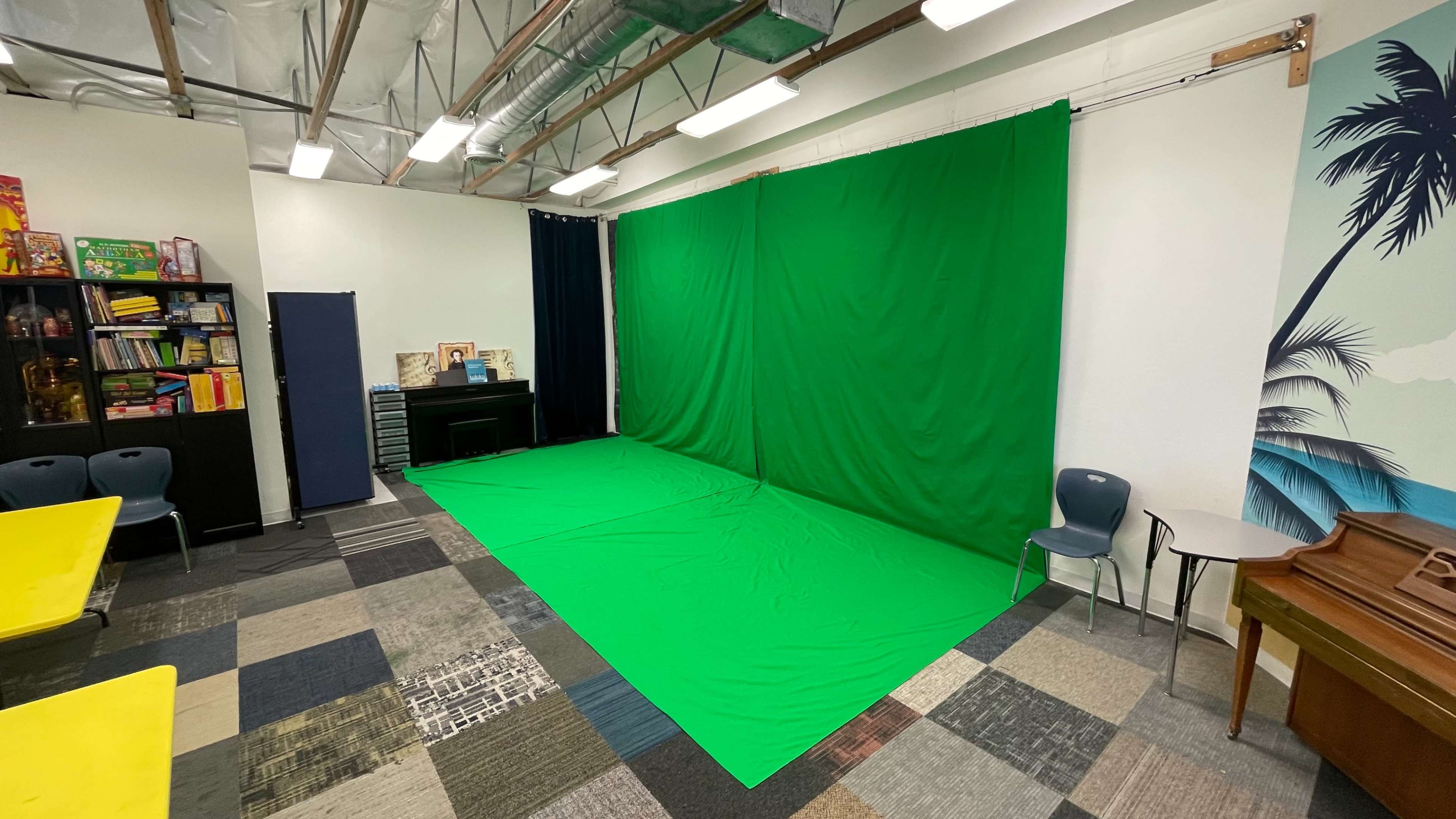 A room with a green screen setup against one wall, surrounded by colorful flooring and furniture, including a piano and storage units.