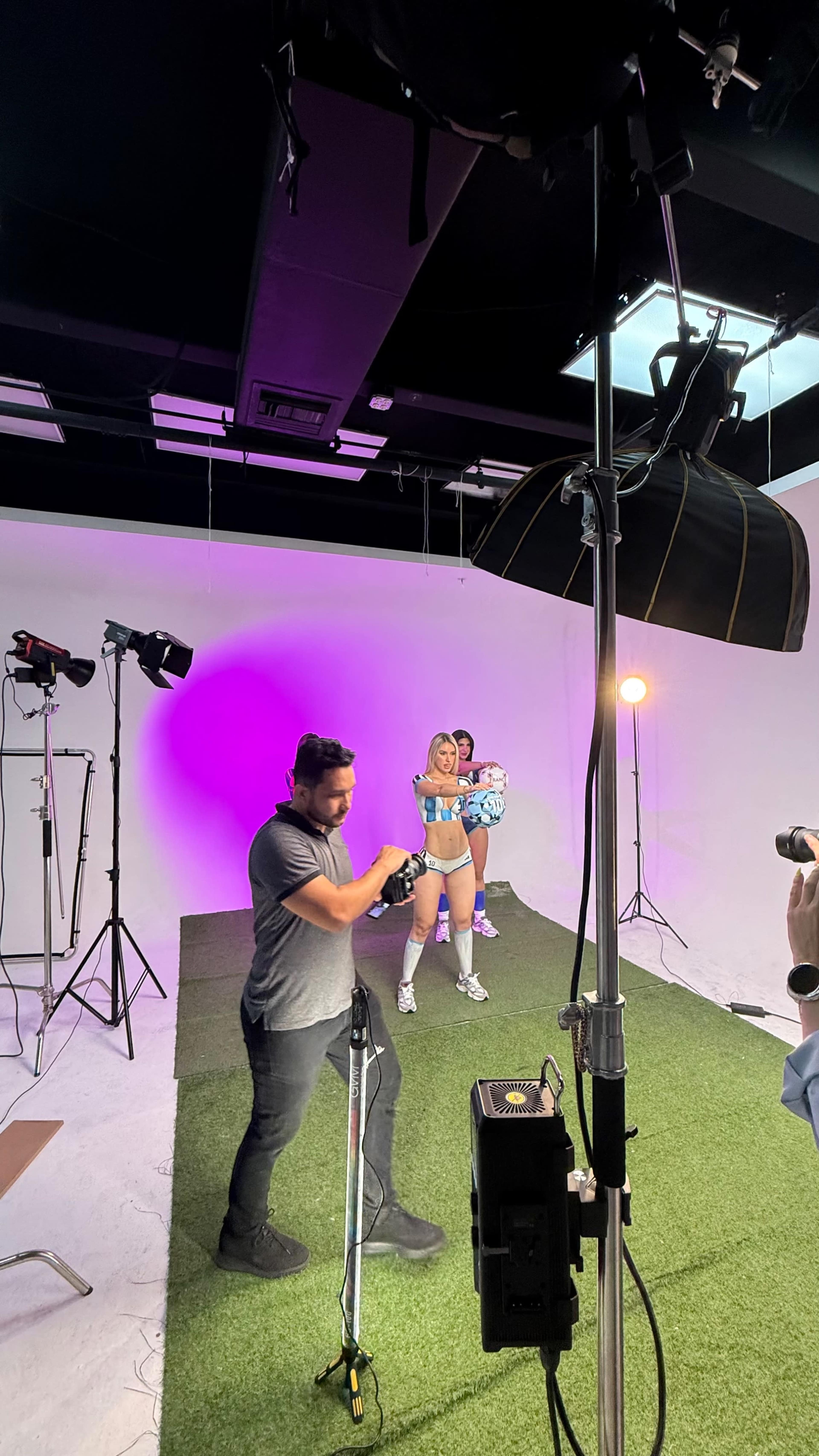 A photographer captures images of two models posing in a studio set with green flooring and bright lights.