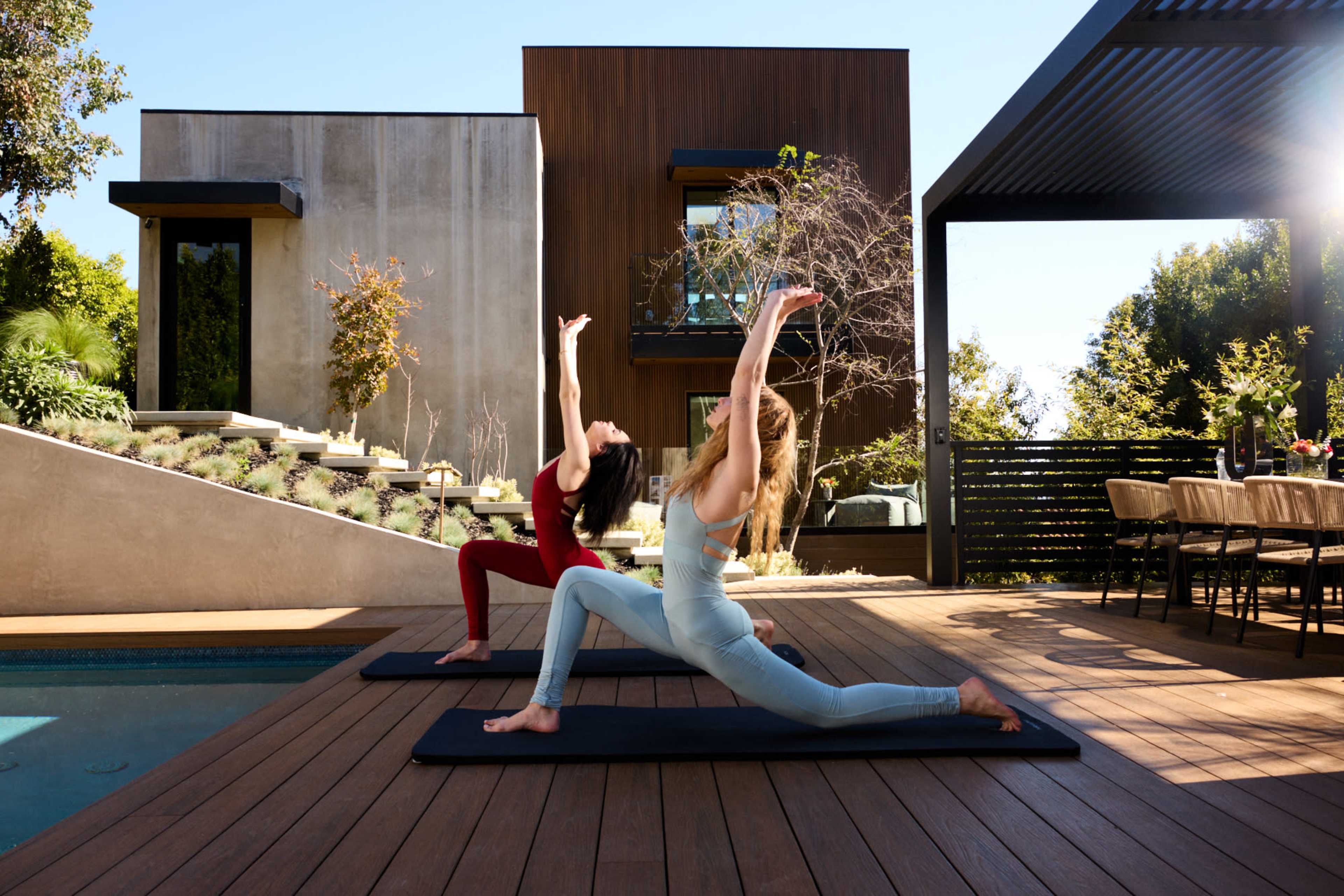 Two women perform yoga poses on mats beside a swimming pool in a modern outdoor setting.