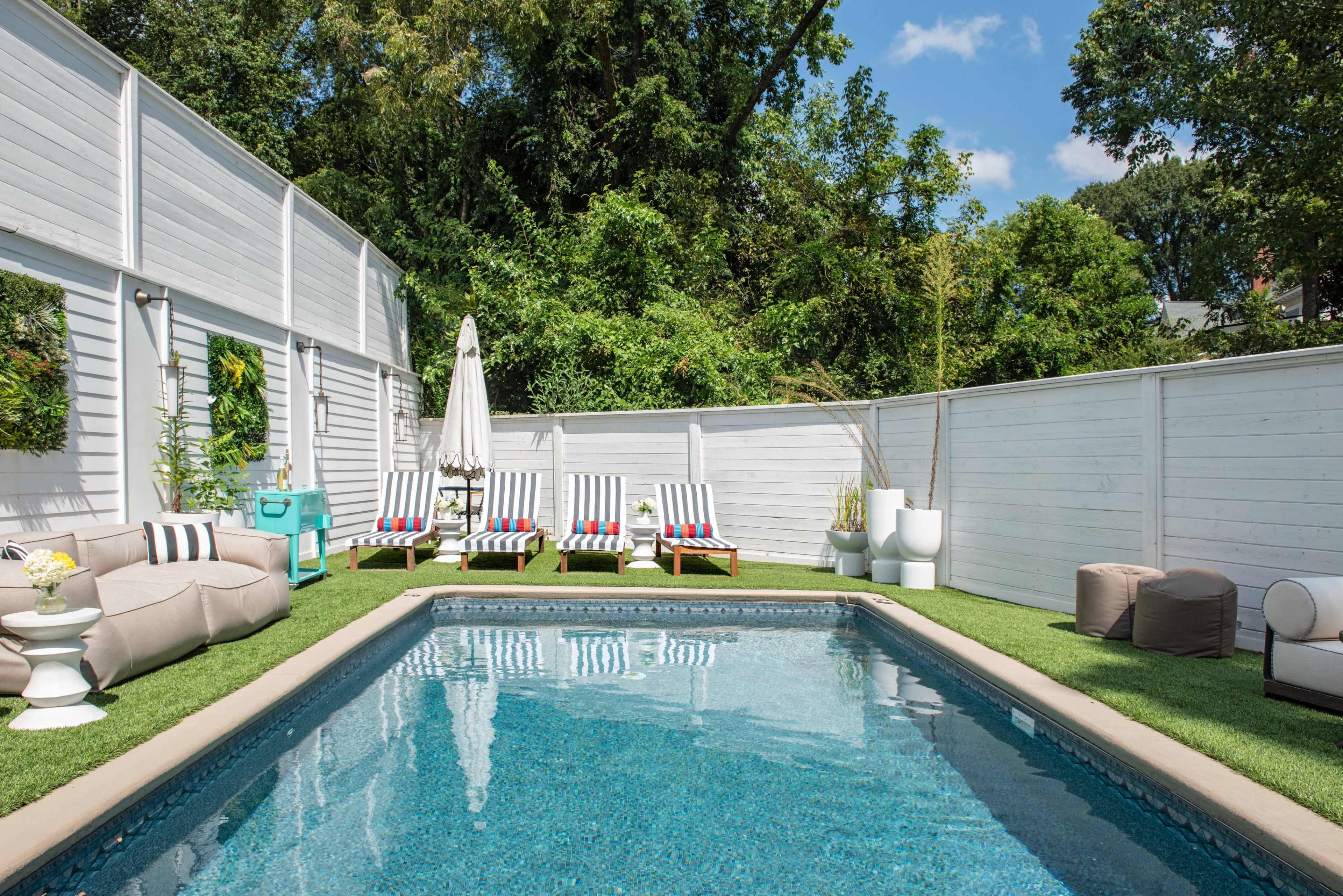 A swimming pool surrounded by lounge chairs and a neatly landscaped outdoor area with greenery and a white privacy fence.