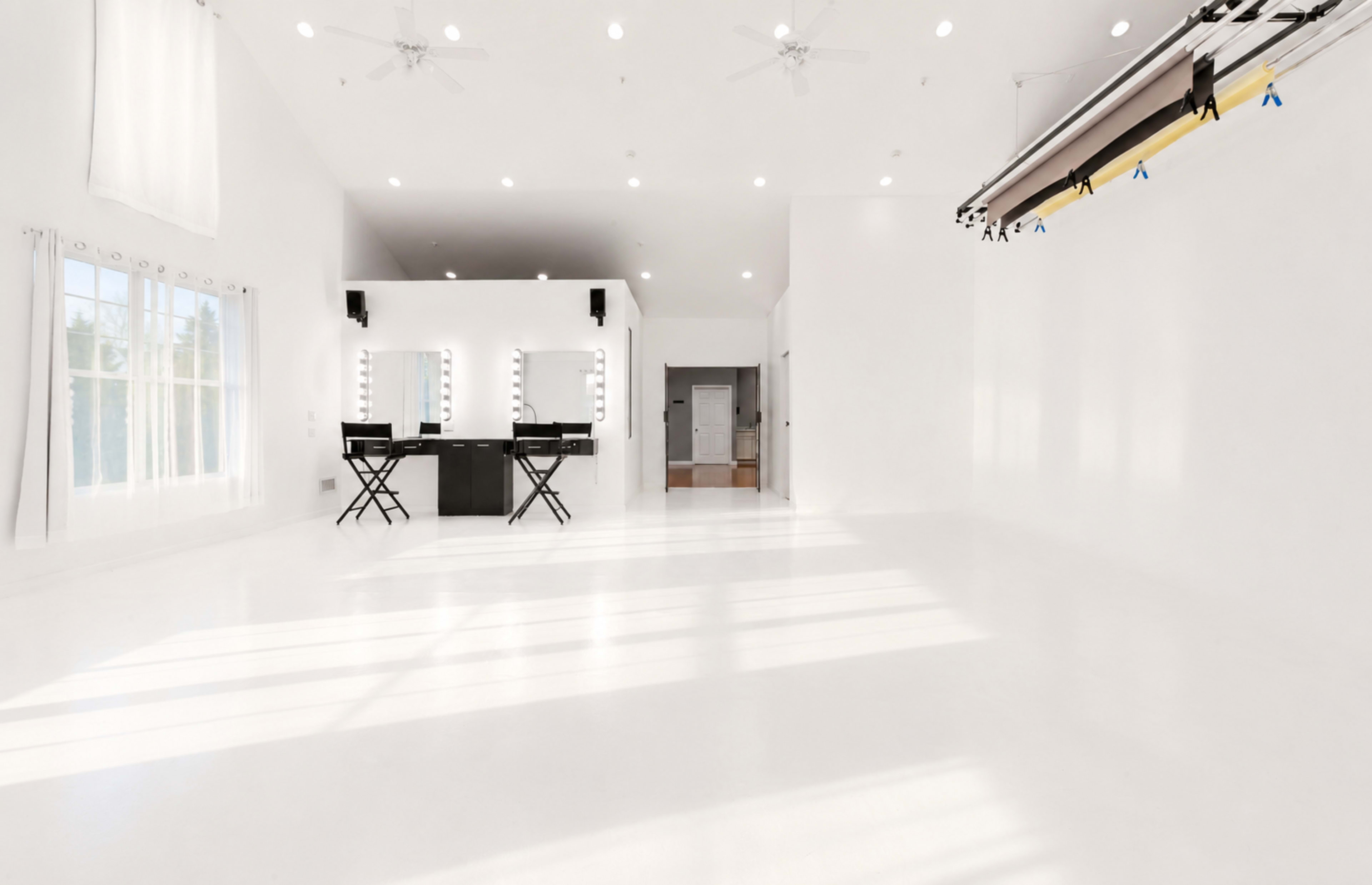 The image shows a spacious, well-lit studio with white walls, a polished floor, and two black chairs facing a vanity with mirrors.