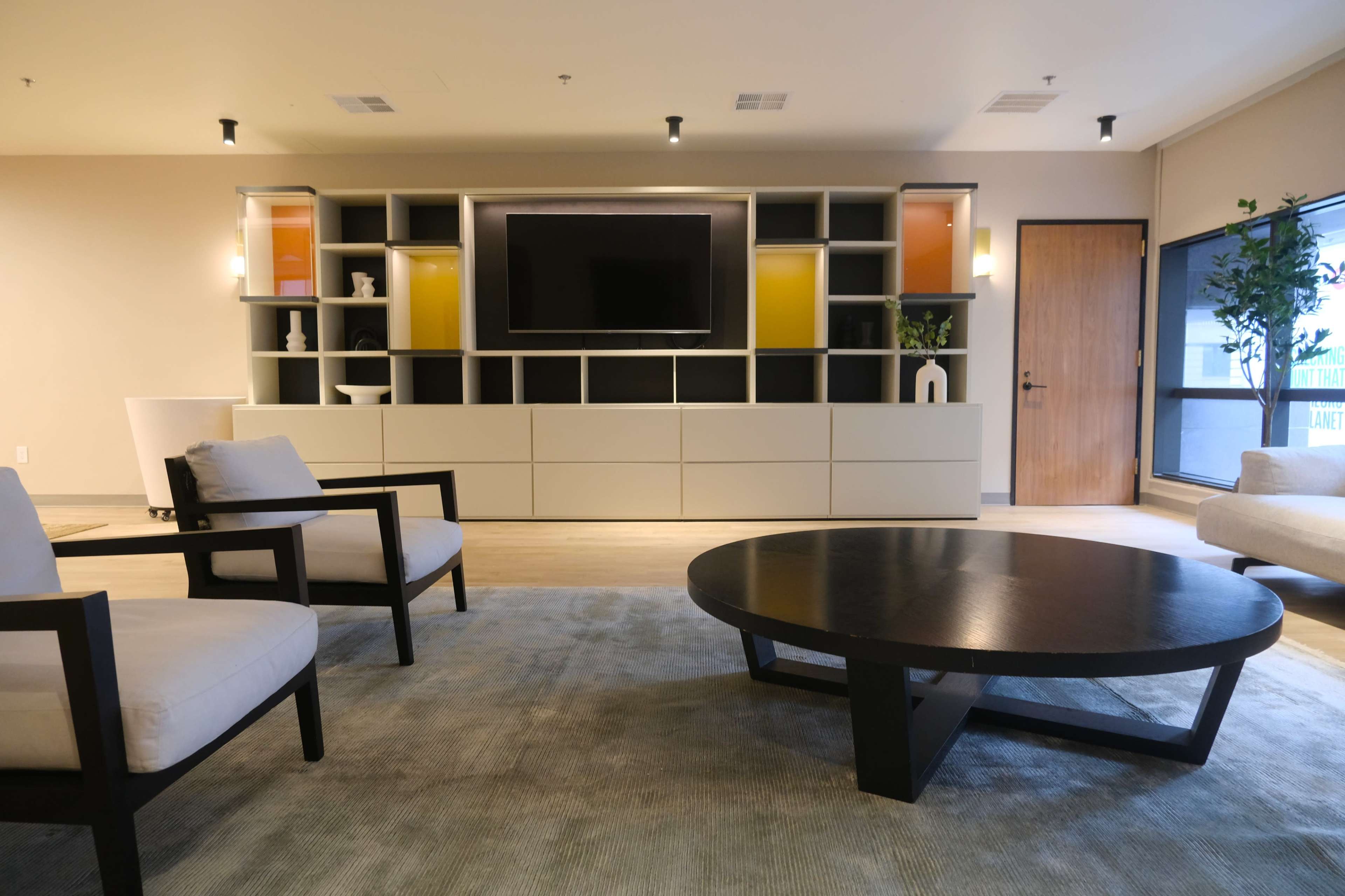 A modern living room with two armchairs, a round black coffee table, and a wall-mounted television set against a light-colored cabinetry.