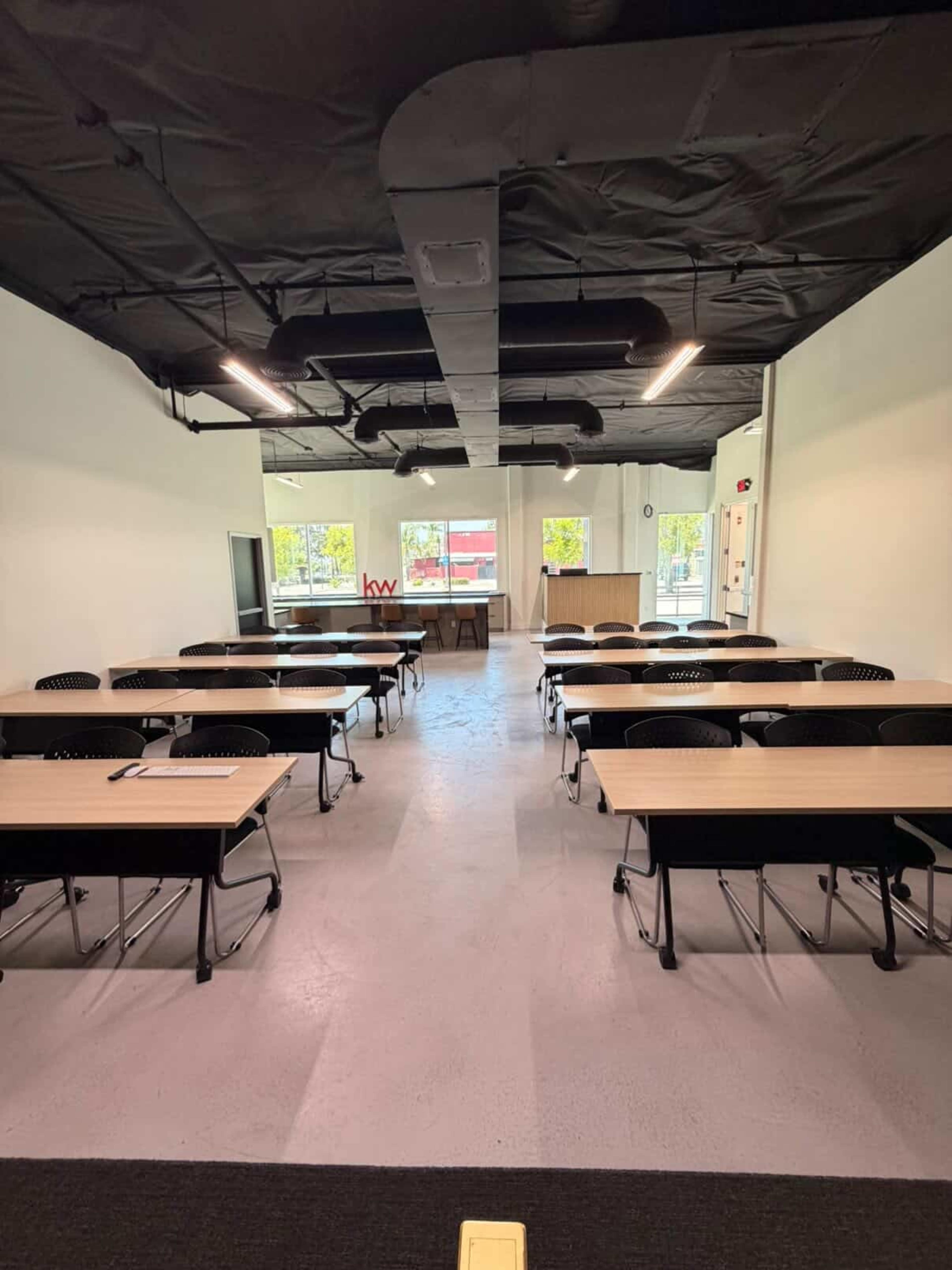 The image shows a classroom with rows of tables and chairs, large windows, and a view of an exterior space.
