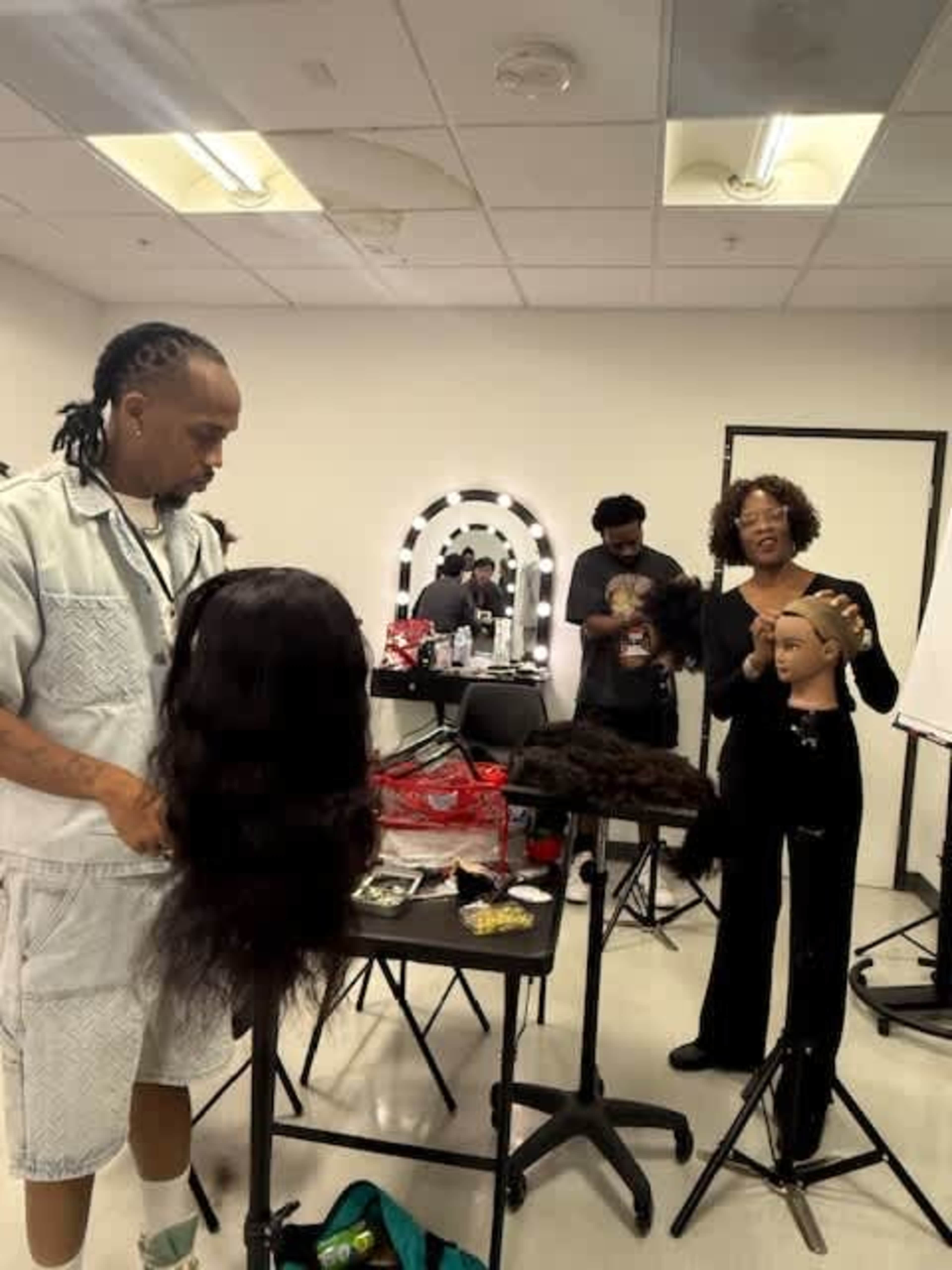 A hairstyling class is in session with two instructors demonstrating techniques on mannequin heads while students observe.