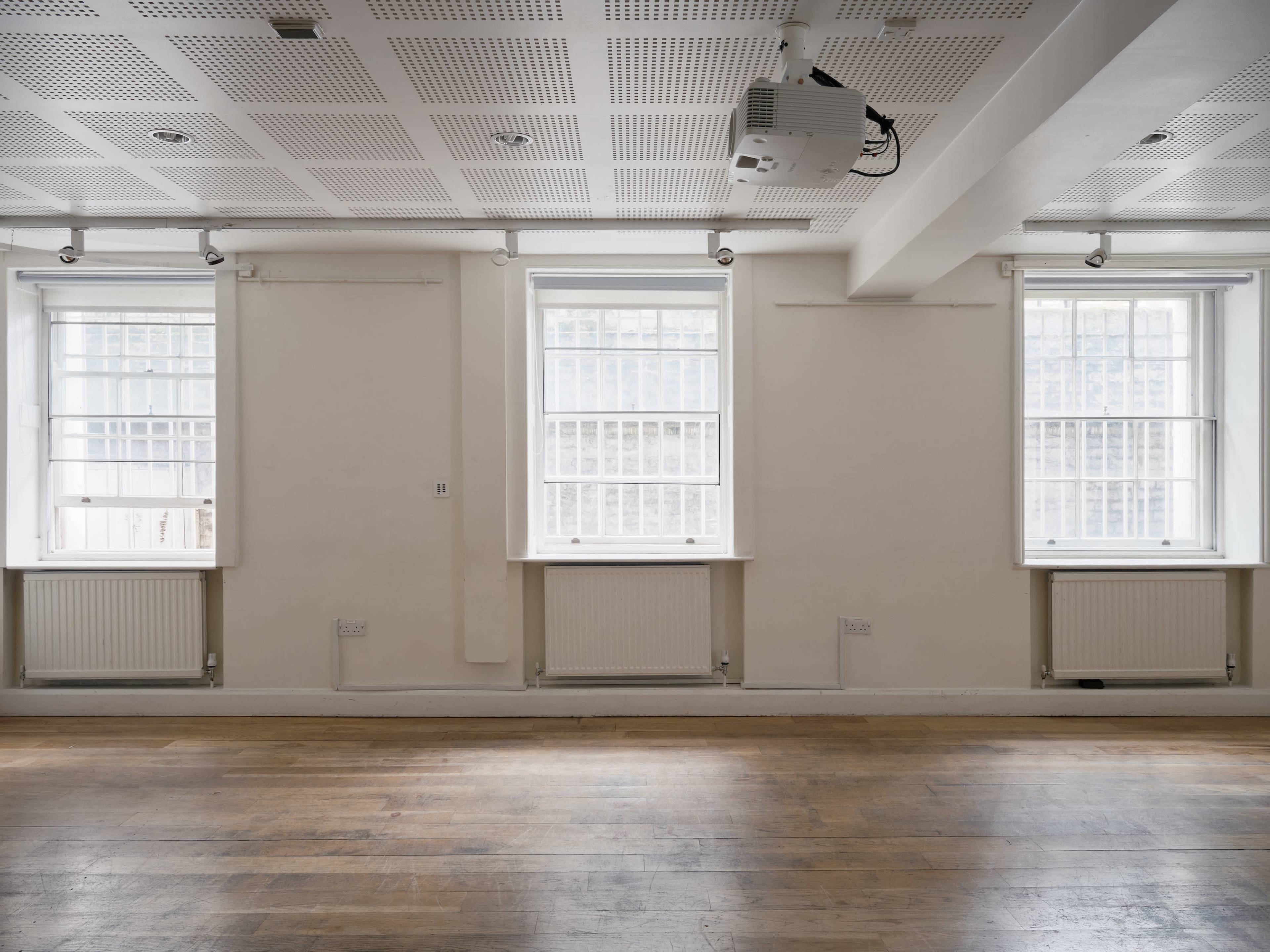 The image shows an empty room with wooden flooring, featuring three windows along one wall and a ceiling-mounted projector.