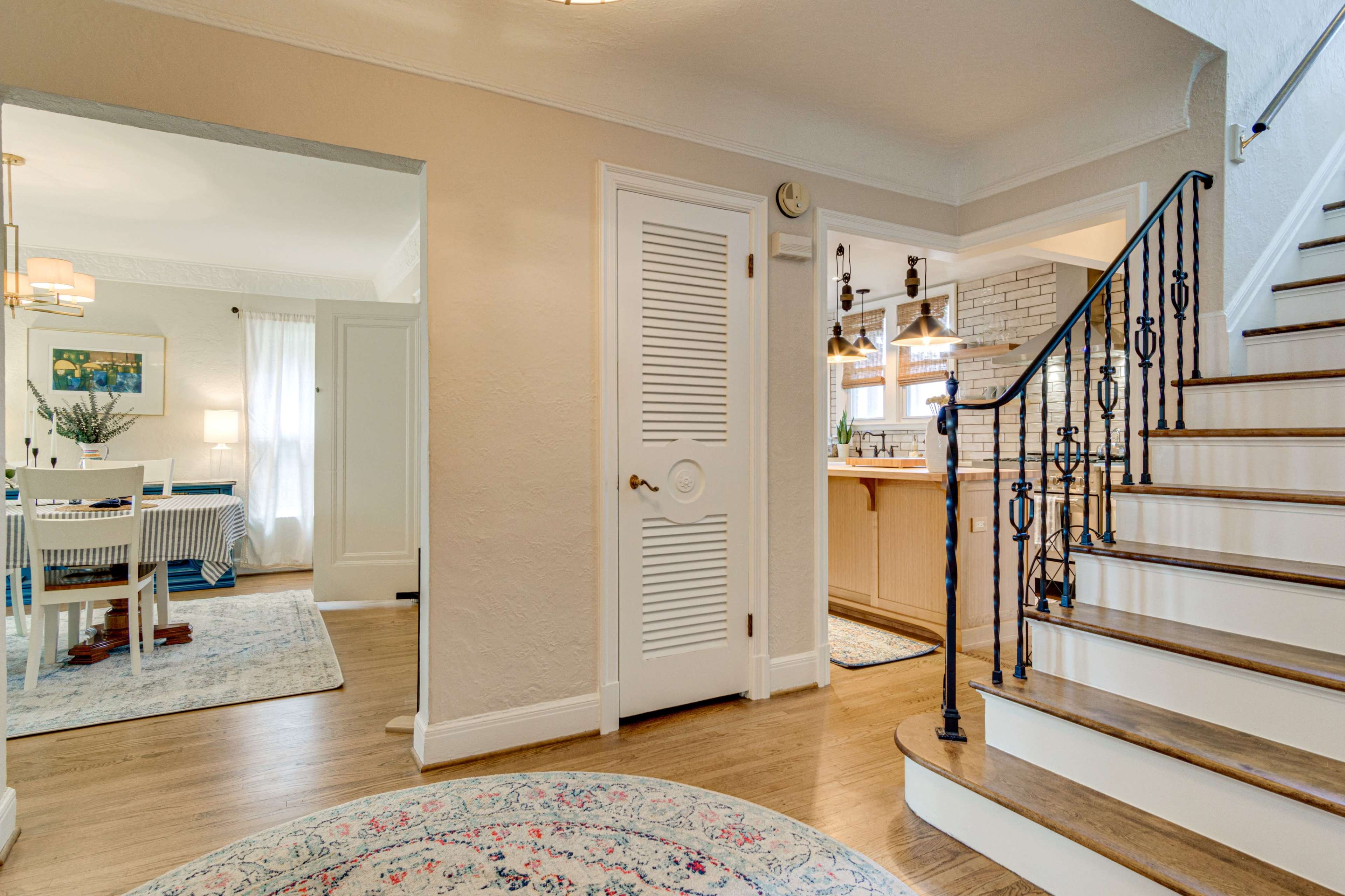The image shows a well-lit entryway with a staircase on the right, leading to a kitchen visible through an open doorway, while a dining area is seen to the left.