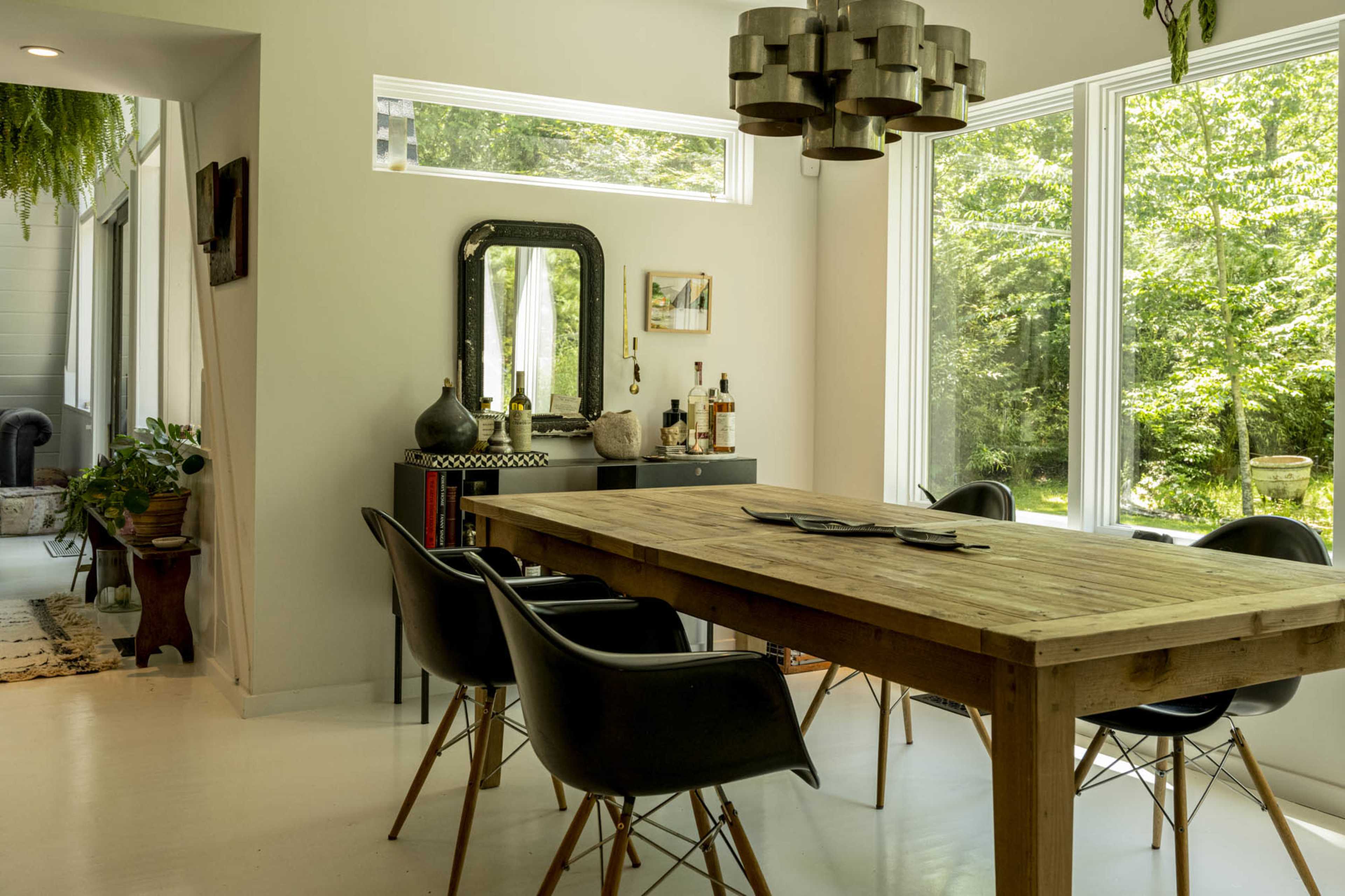 The image depicts a modern dining area featuring a wooden table surrounded by black chairs, with large windows offering a view of greenery outside.