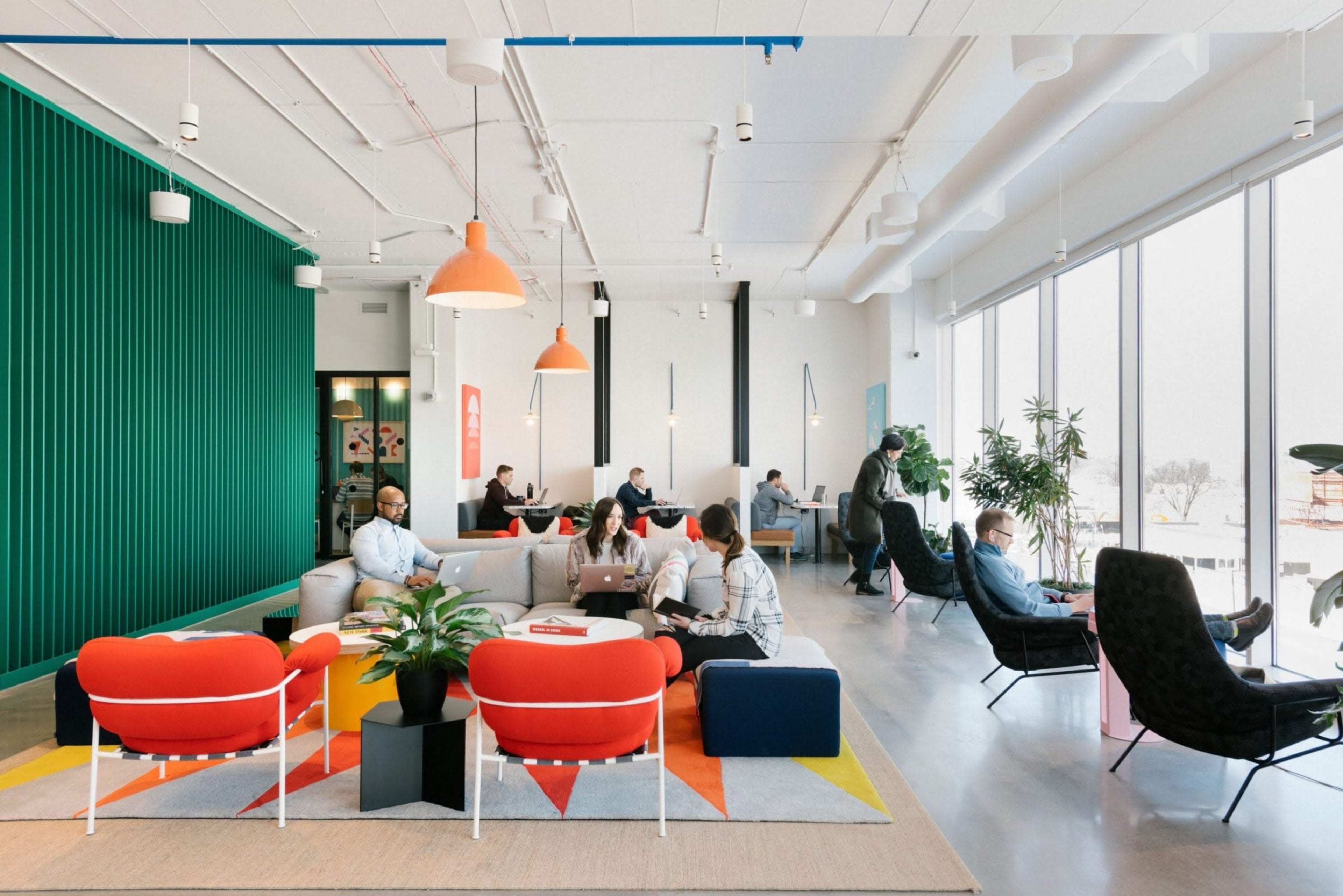 A modern office lounge area with colorful seating, large windows, and several people working and socializing.