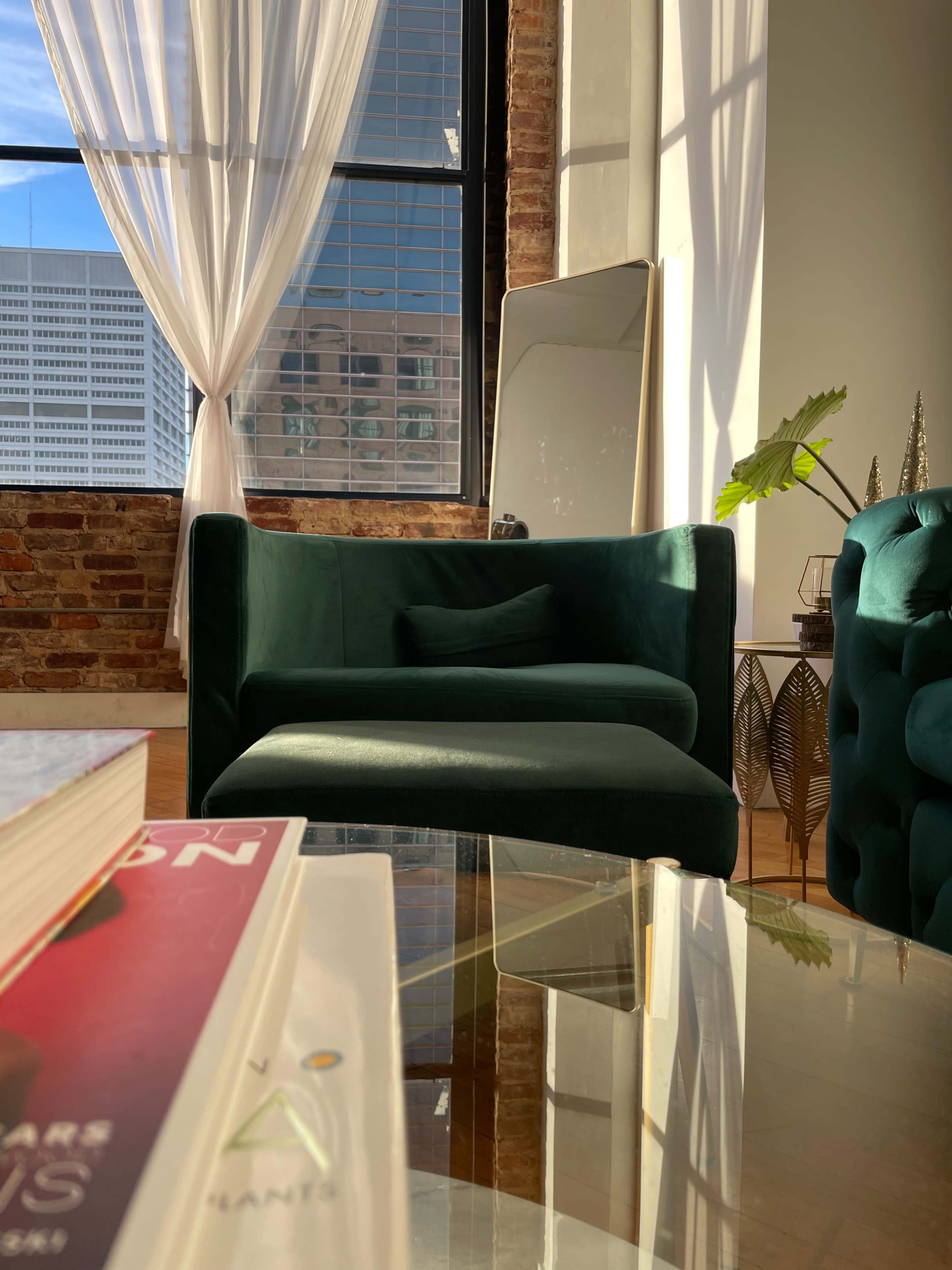 A green armchair and a mirrored table are positioned near a large window with sheer curtains, allowing sunlight to illuminate the room.