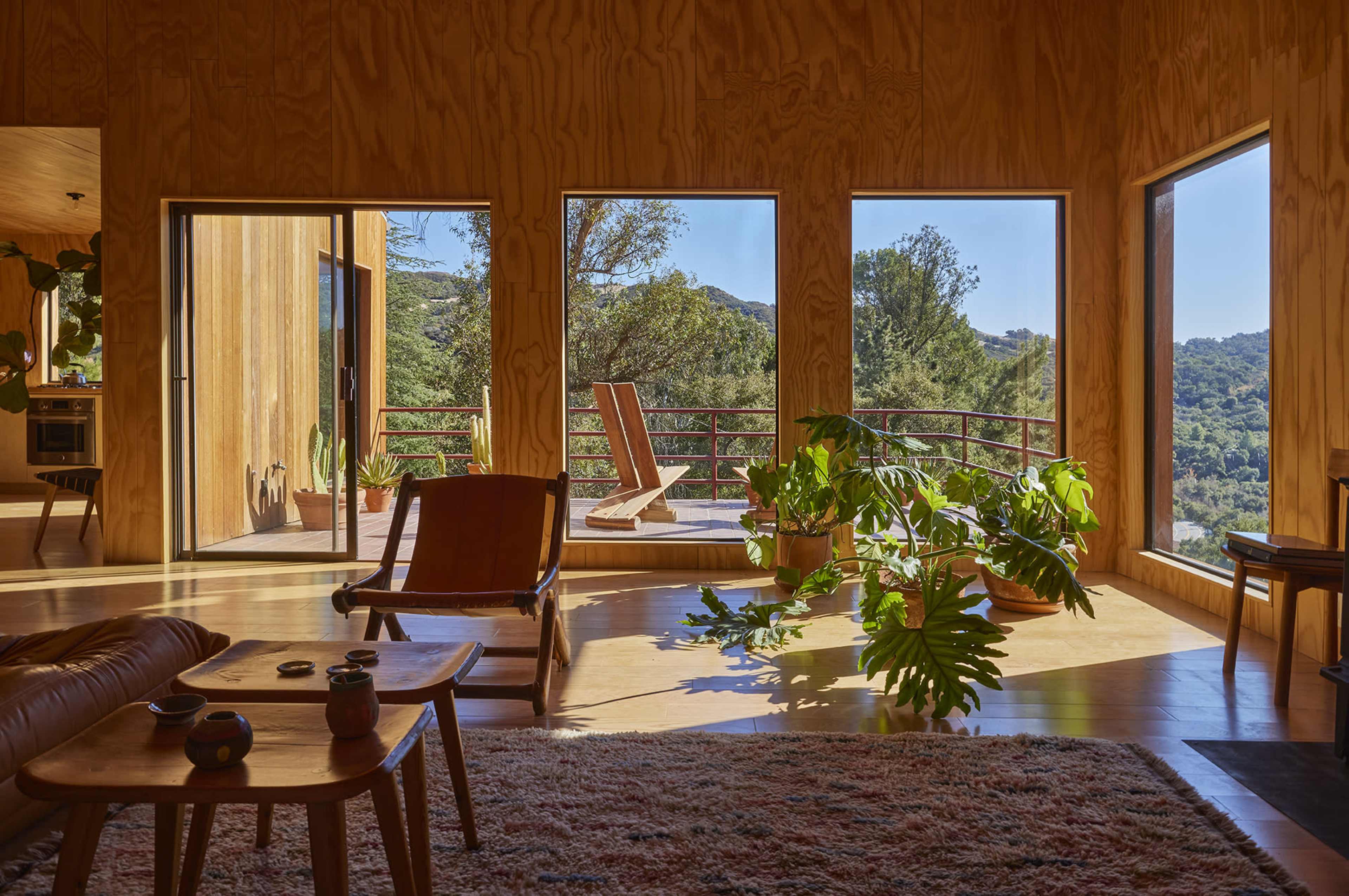 The interior of a wooden house features large windows with a view of greenery, a lounge chair, and potted plants.