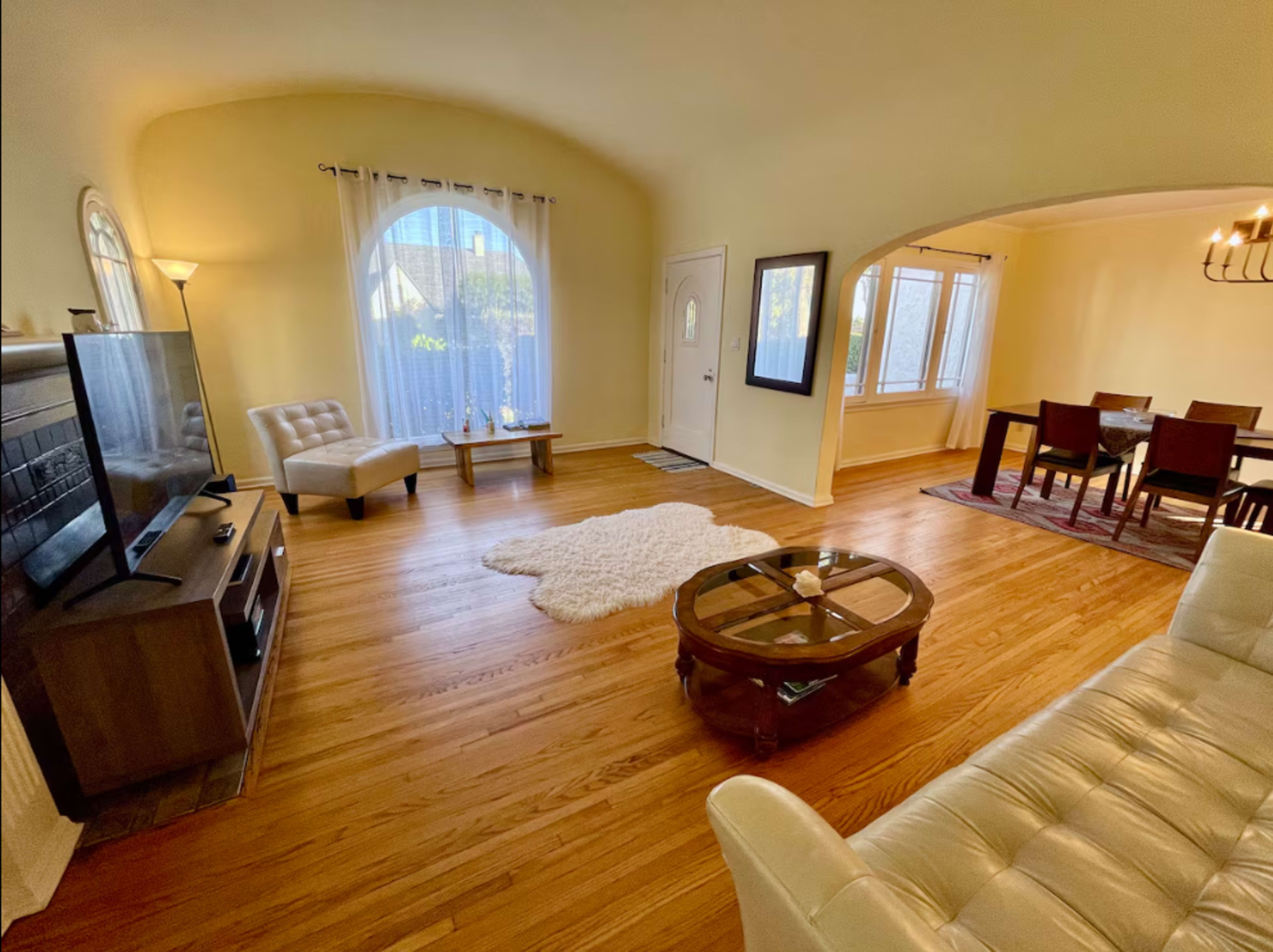 The image shows a bright living room with hardwood floors, a television, a dining area, and large windows allowing natural light in.