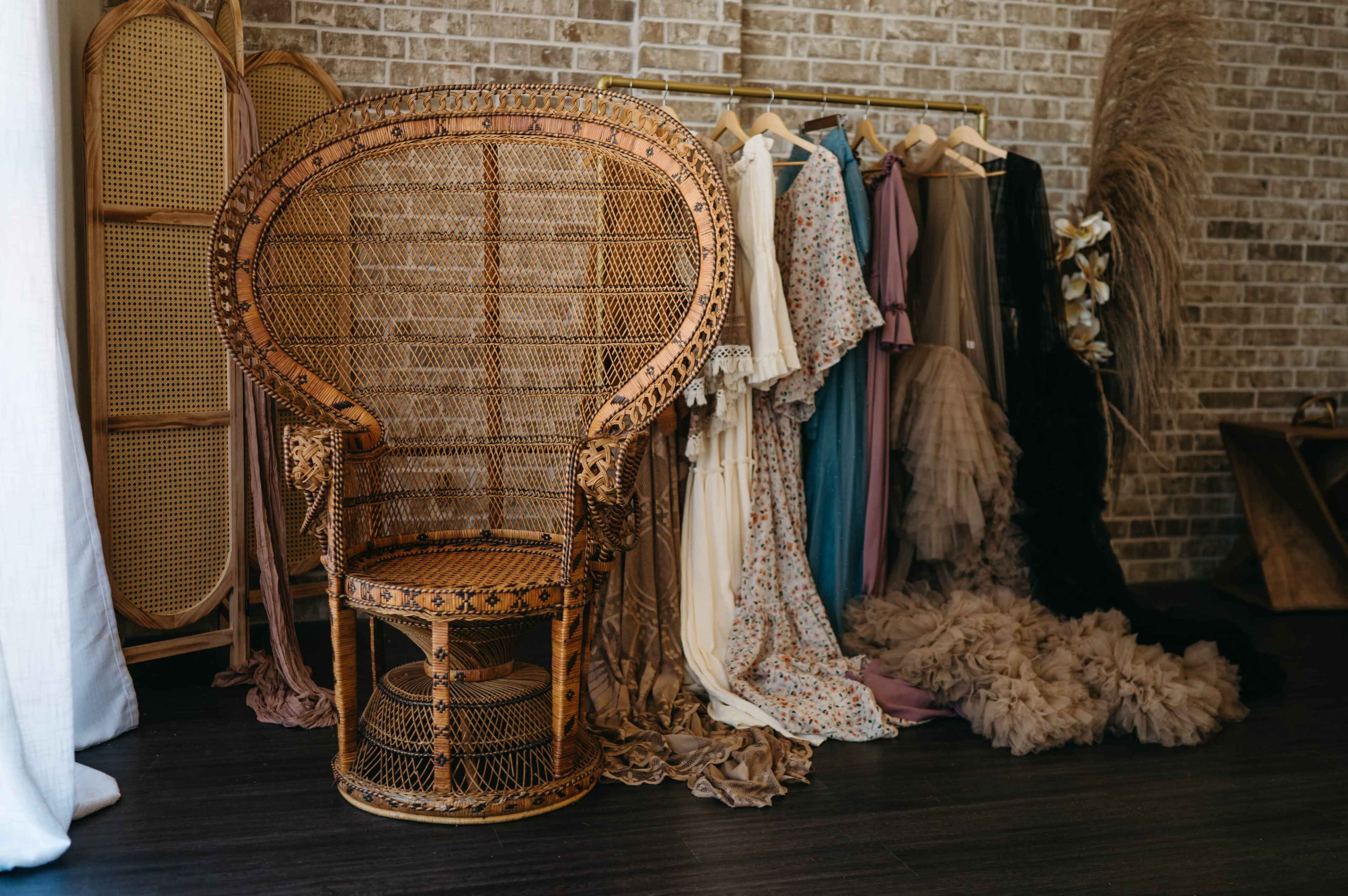 A large, intricately woven rattan chair stands next to a clothing rack displaying various elegant dresses against a brick wall.