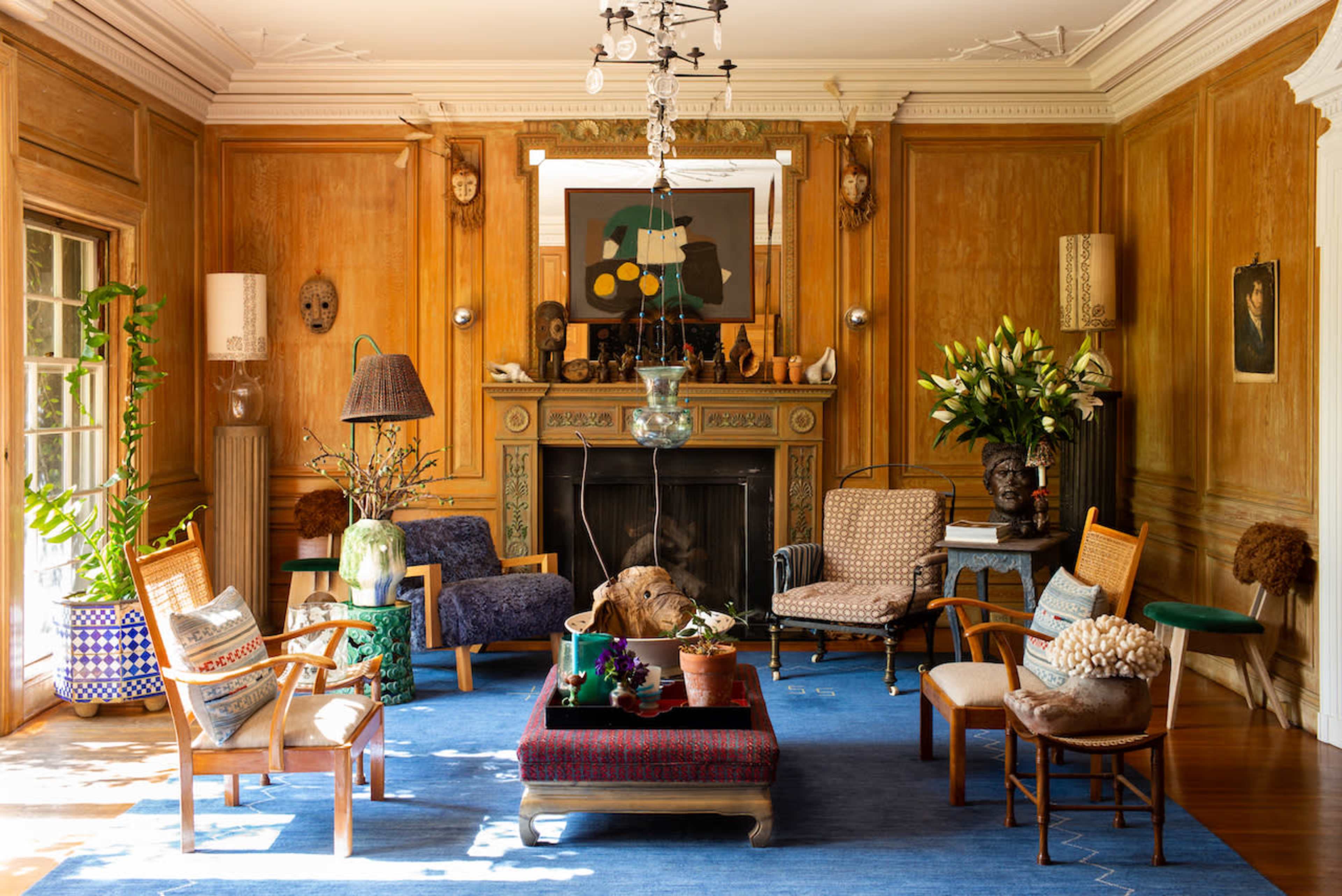 The image shows a living room with wooden walls, a fireplace, various seating arrangements, and decorative plants.