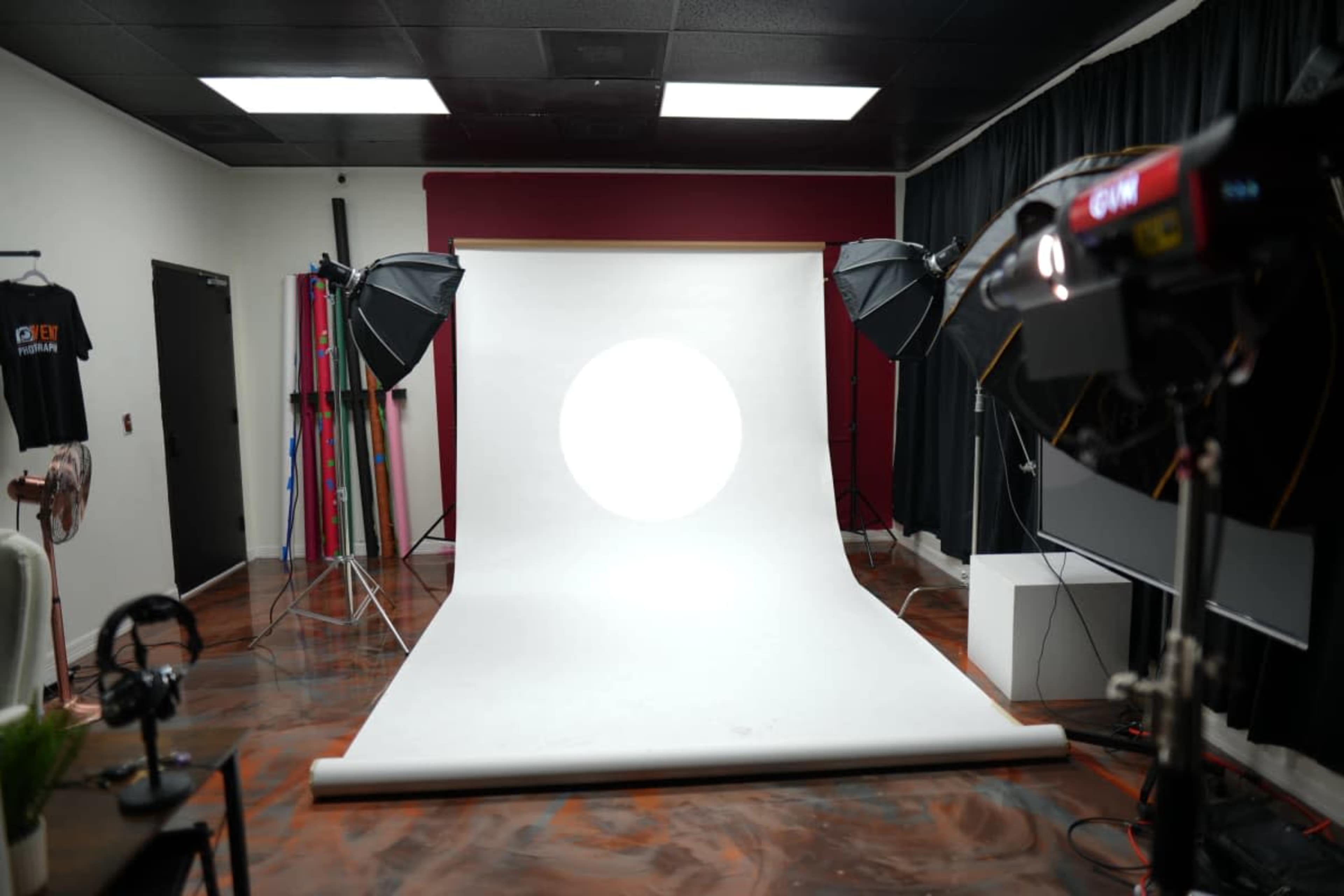 The image shows a photography studio with a white backdrop, two softbox lights, and various equipment arranged around the space.