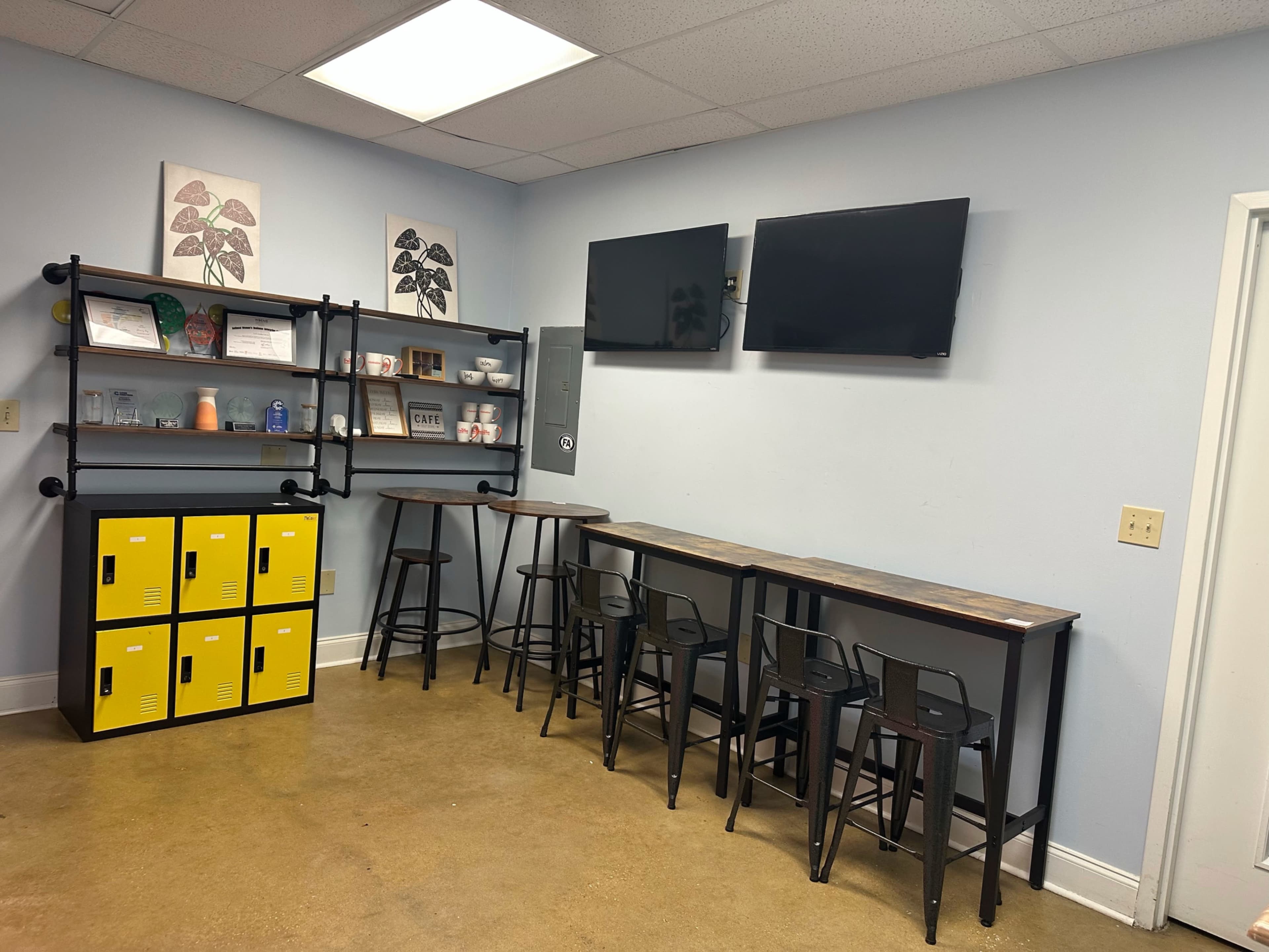The image shows a small break room with two large tables, black stools, a wall shelf displaying various items, and two mounted televisions.