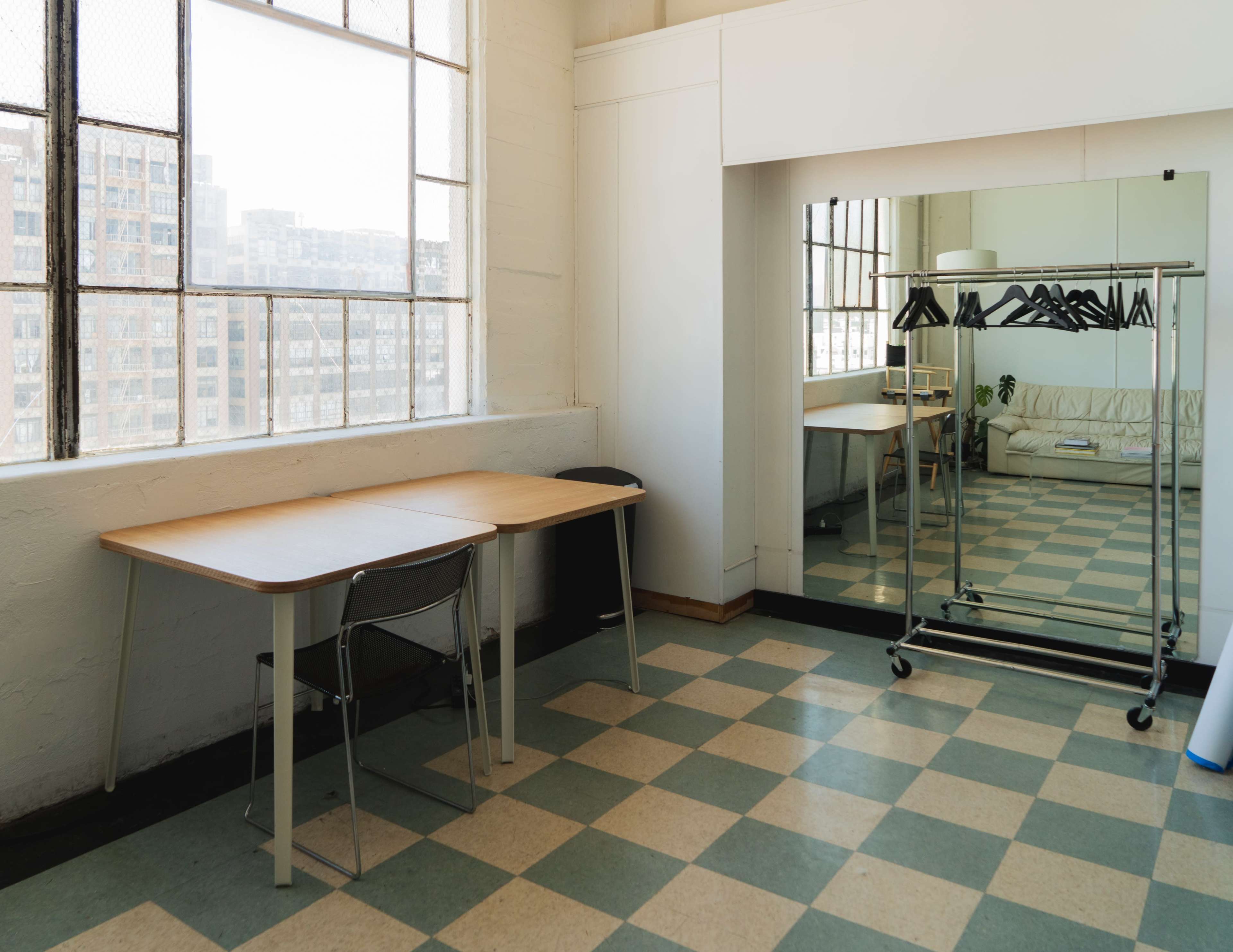 A bright room features a wooden table with a chair, a mirror, and a clothing rack with hangers against a backdrop of large windows.