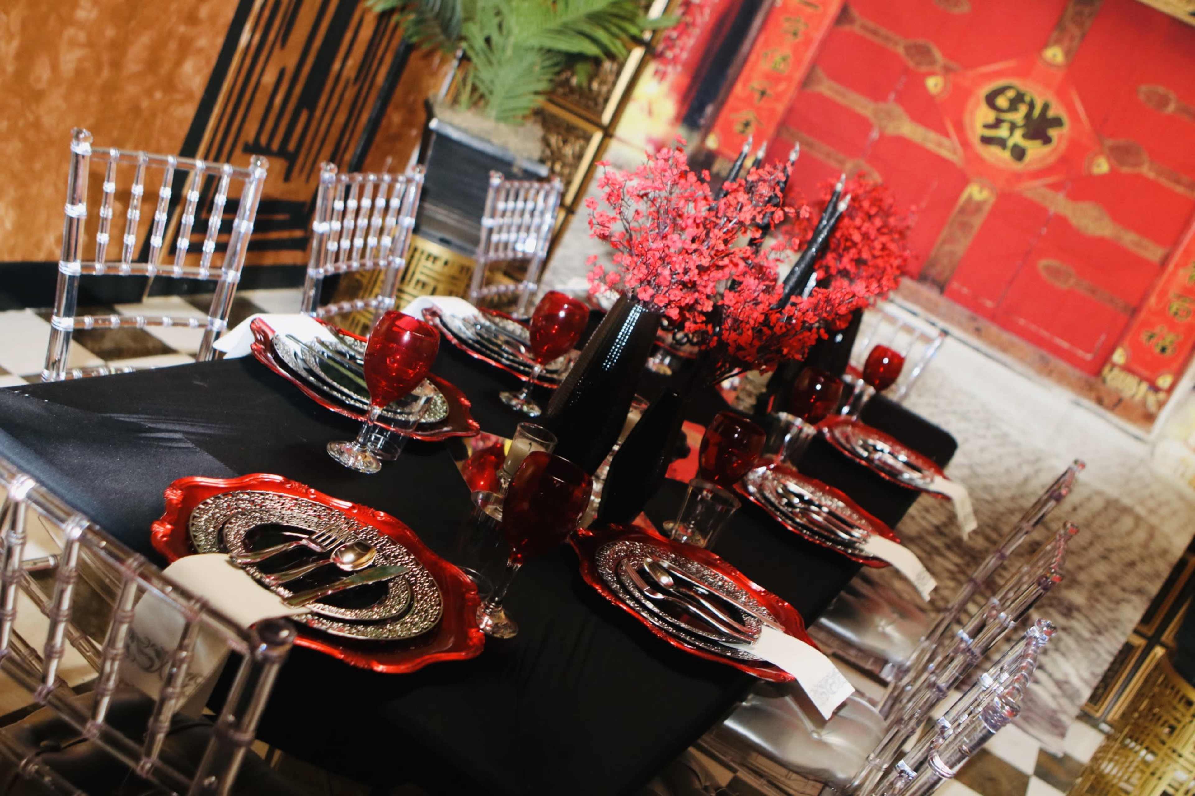 A dining table is set with ornate dishware and red floral arrangements against a backdrop of a red door with traditional motifs.